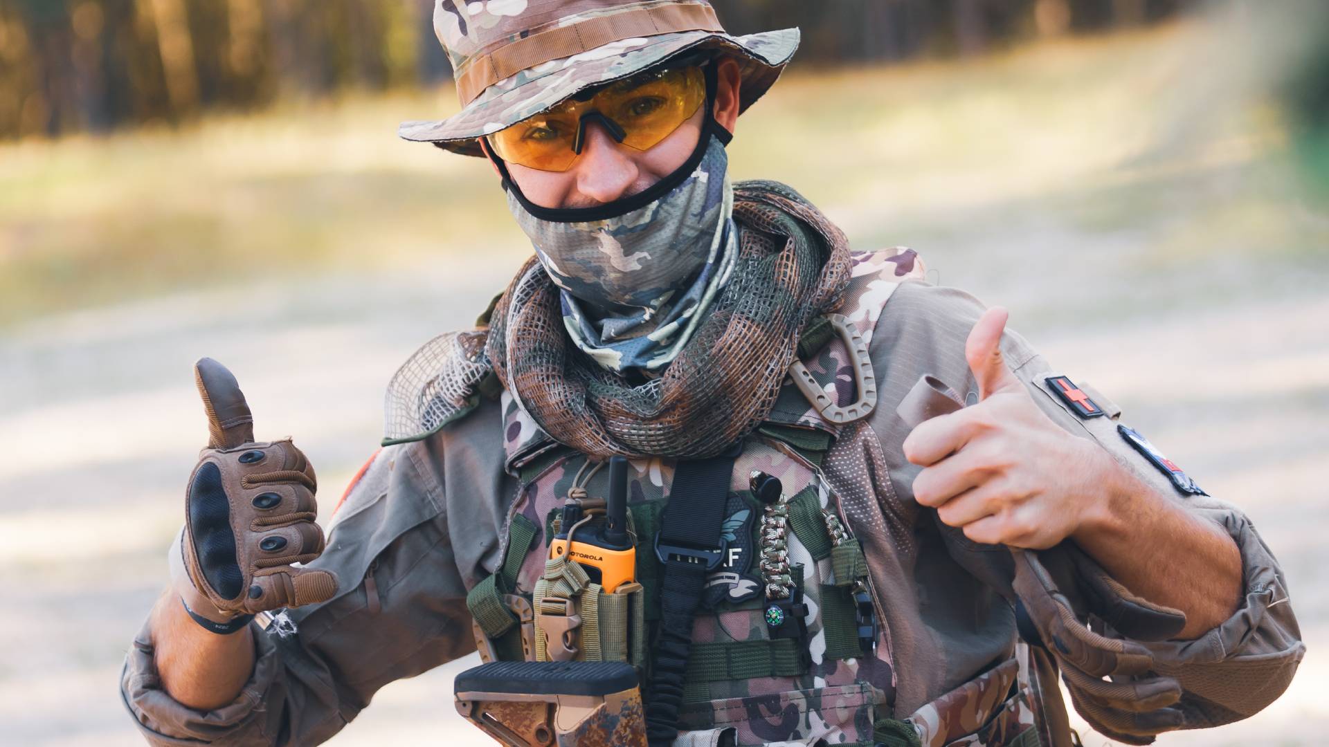Airsoft-Spieler in Tarnkleidung mit Ausrüstung zeigt Daumen hoch und trägt Schutzbrille.