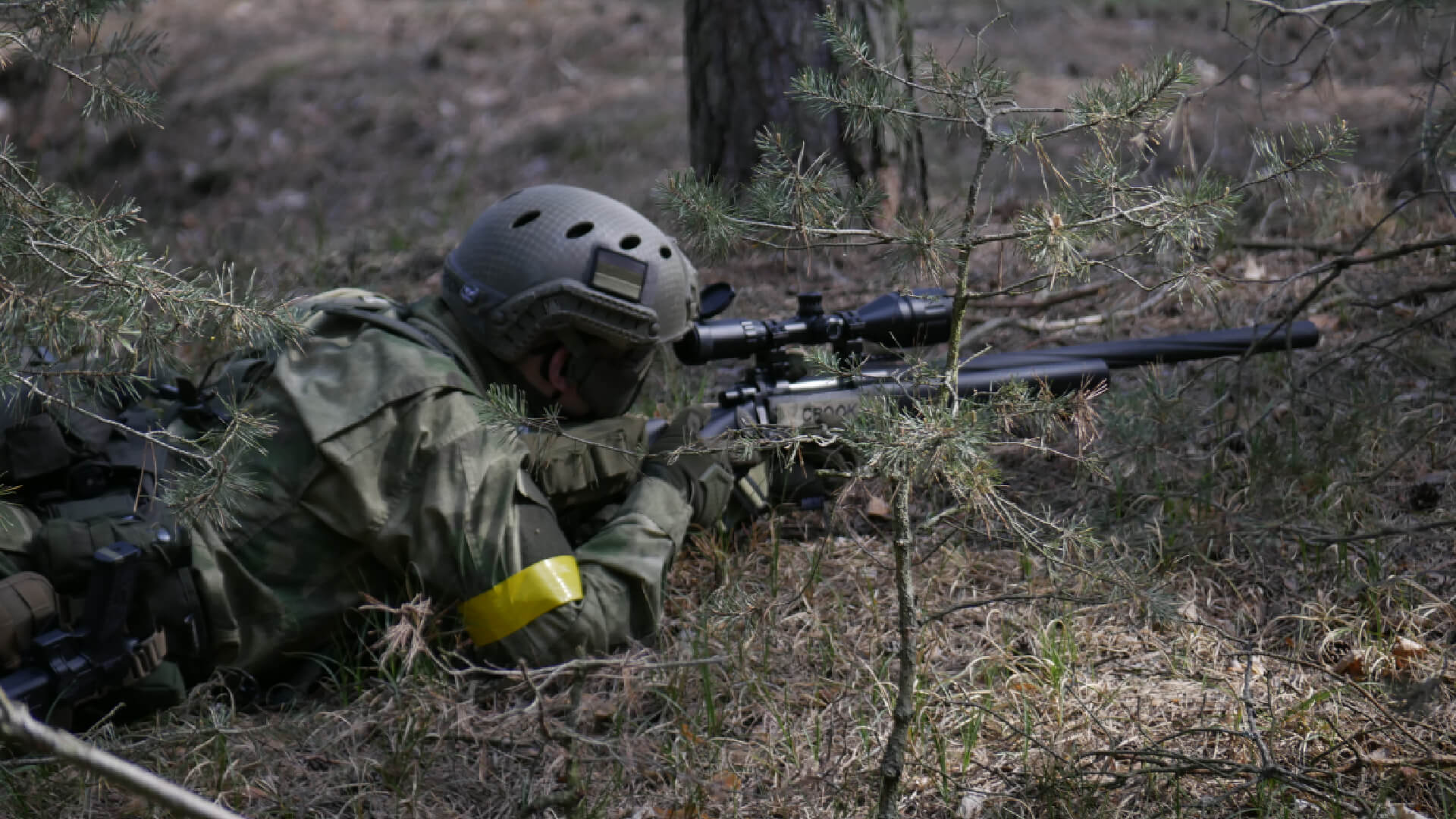 Airsoft-Spieler in Tarnkleidung zielt mit Gewehr im Wald, gelbes Armband am Arm.