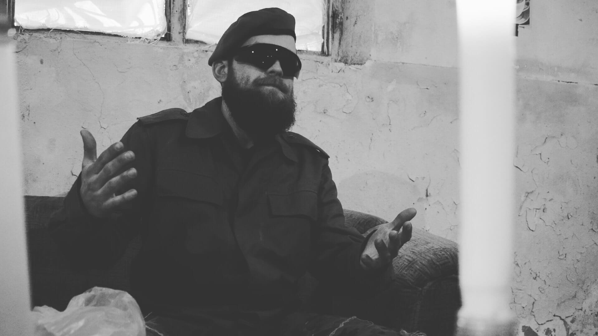 Person im Tarnanzug mit Barett und Sonnenbrille sitzt in einem alten Raum, gestikuliert. Schwarz-Weiß-Foto.