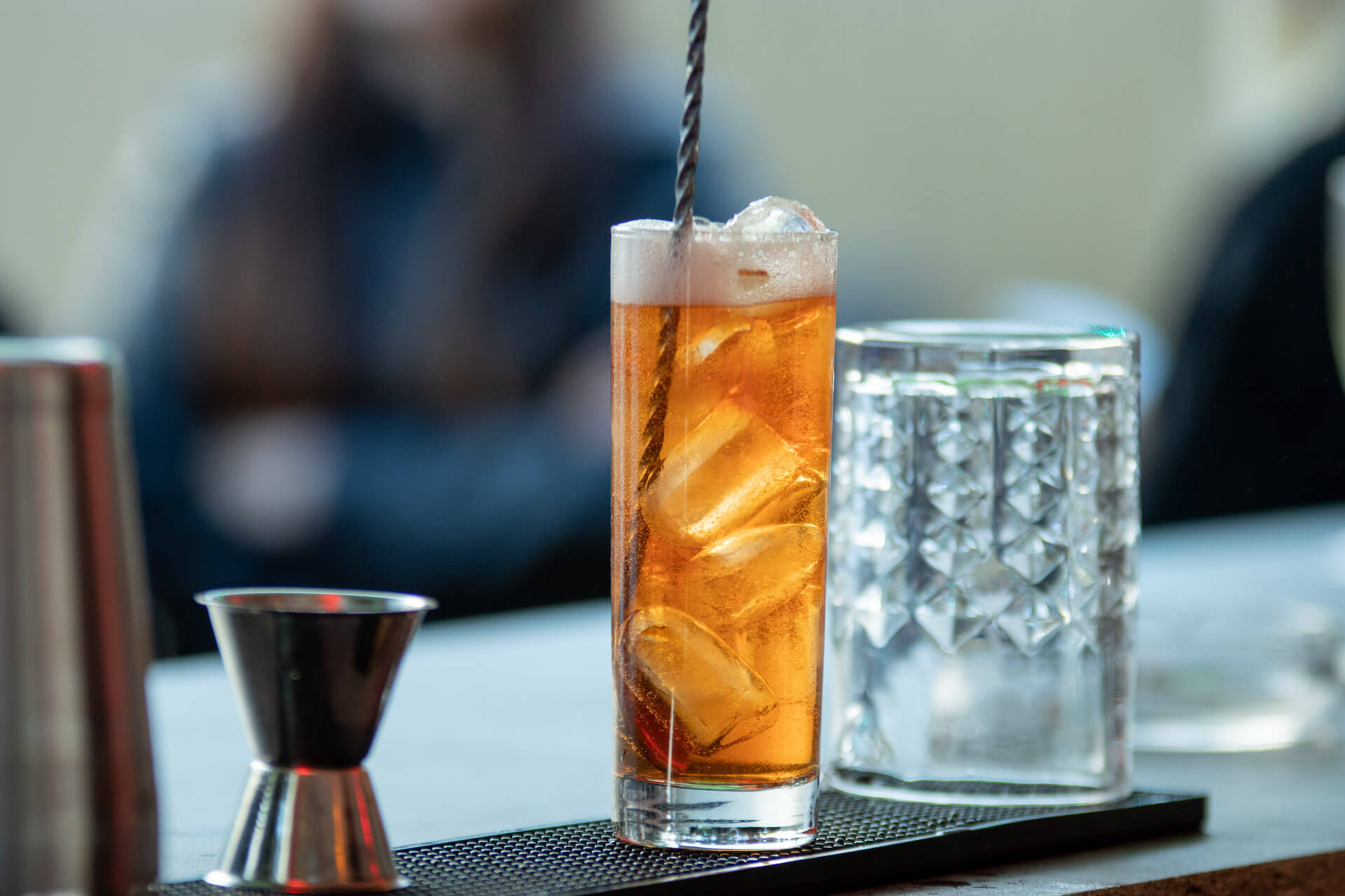 Ein hohes Glas mit einem Eistee-ähnlichen Getränk, gefüllt mit Eiswürfeln und einem Rührstab. Daneben steht ein Messbecher.