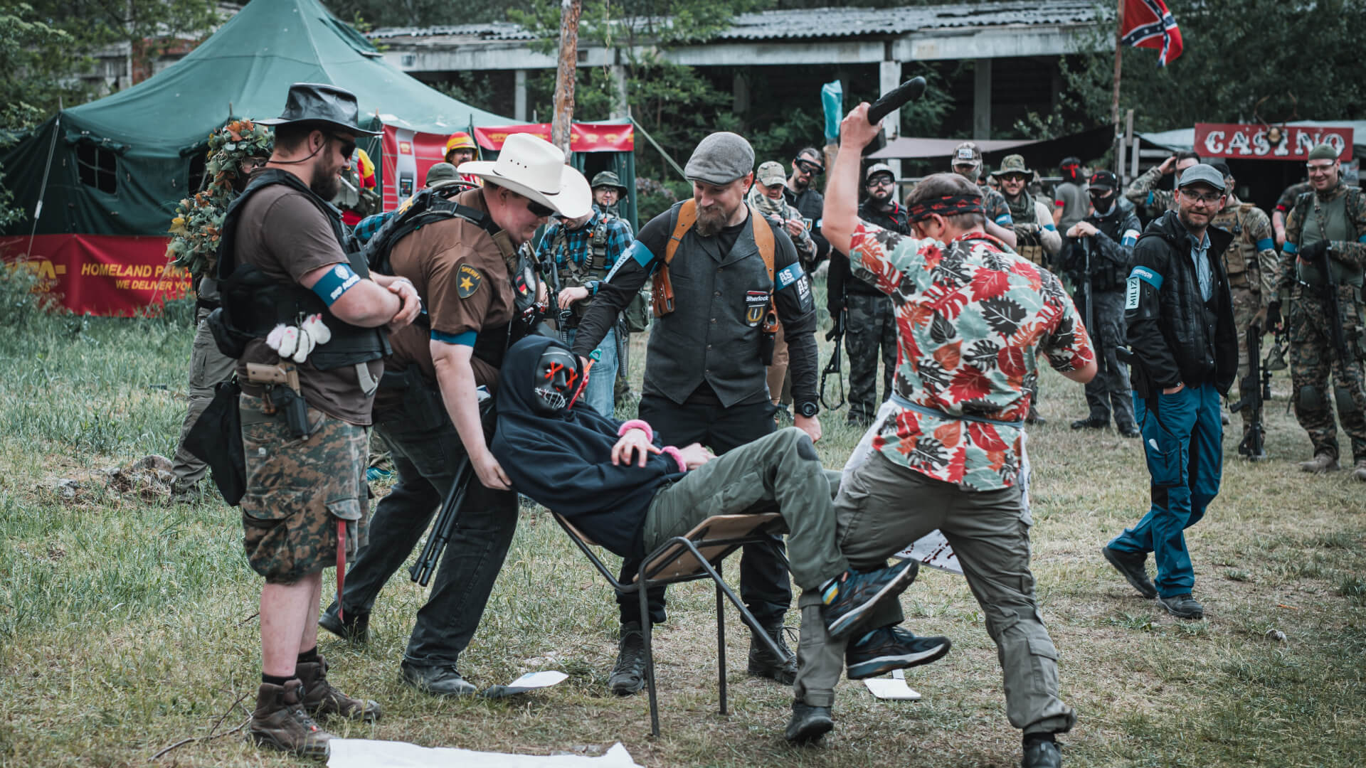 Airsoft-Spieler in bunten Outfits führen eine Szene auf; ein Spieler sitzt gefesselt auf einem Stuhl.