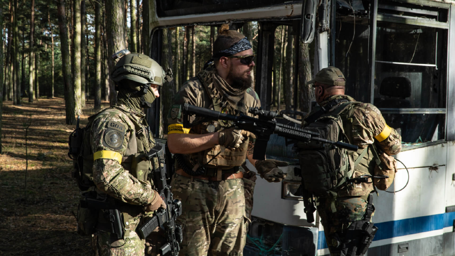 Drei Airsoft-Spieler in Tarnkleidung taktieren neben einem alten Fahrzeug im Wald.