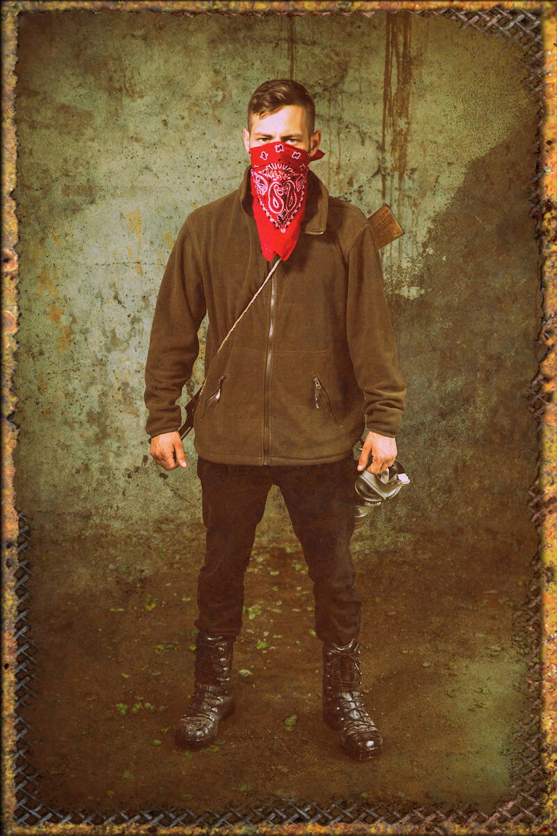 Mann in brauner Jacke und rotem Halstuch mit Gewehr, steht in rauer Umgebung mit Gasmaske.
