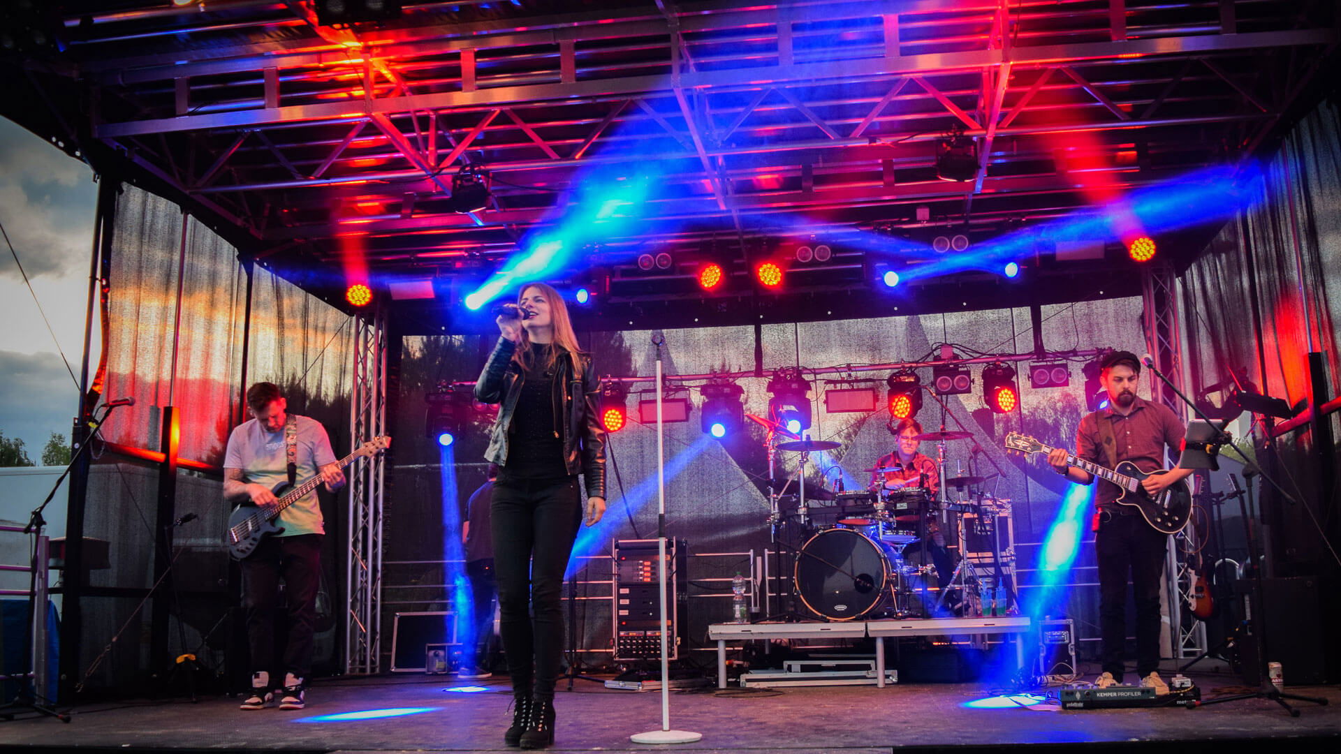 Bandauftritt auf Bühne mit bunter Beleuchtung, Sängerin in der Mitte, Gitarristen und Schlagzeuger im Hintergrund.