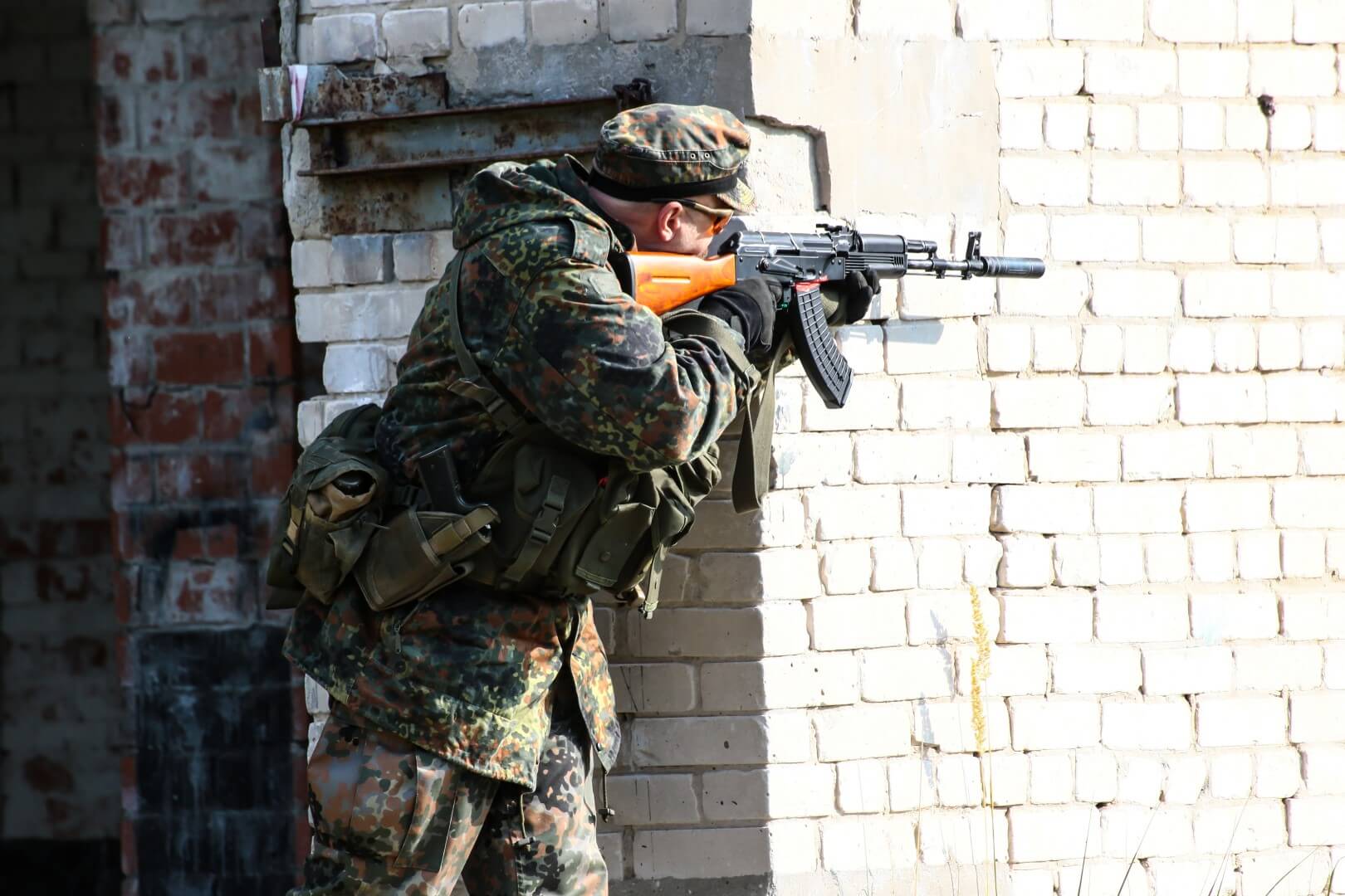 Person in Camouflage, zielt mit Airsoft-Gewehr um eine Ziegelwand.