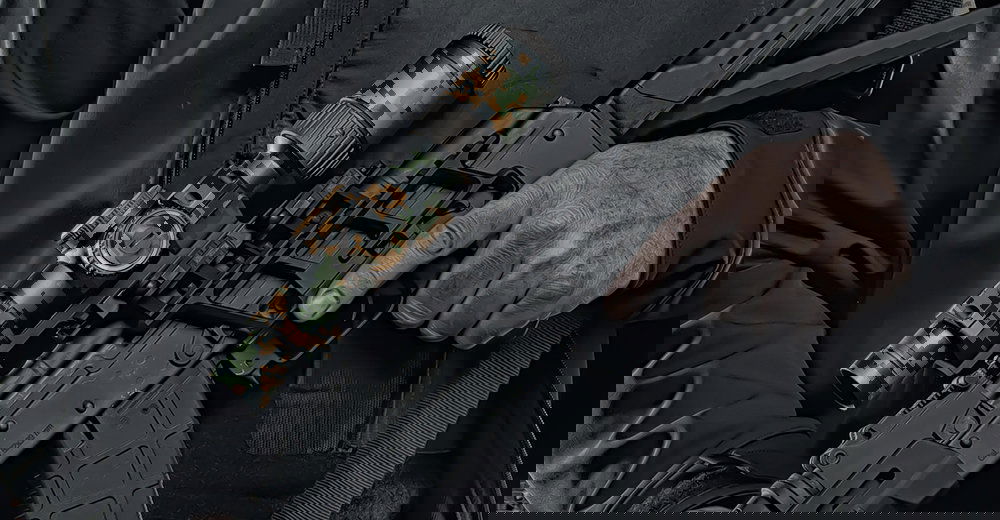 Person hält taktische Airsoft-Waffe mit digital getarntem Zielfernrohr, schwarze Kleidung.