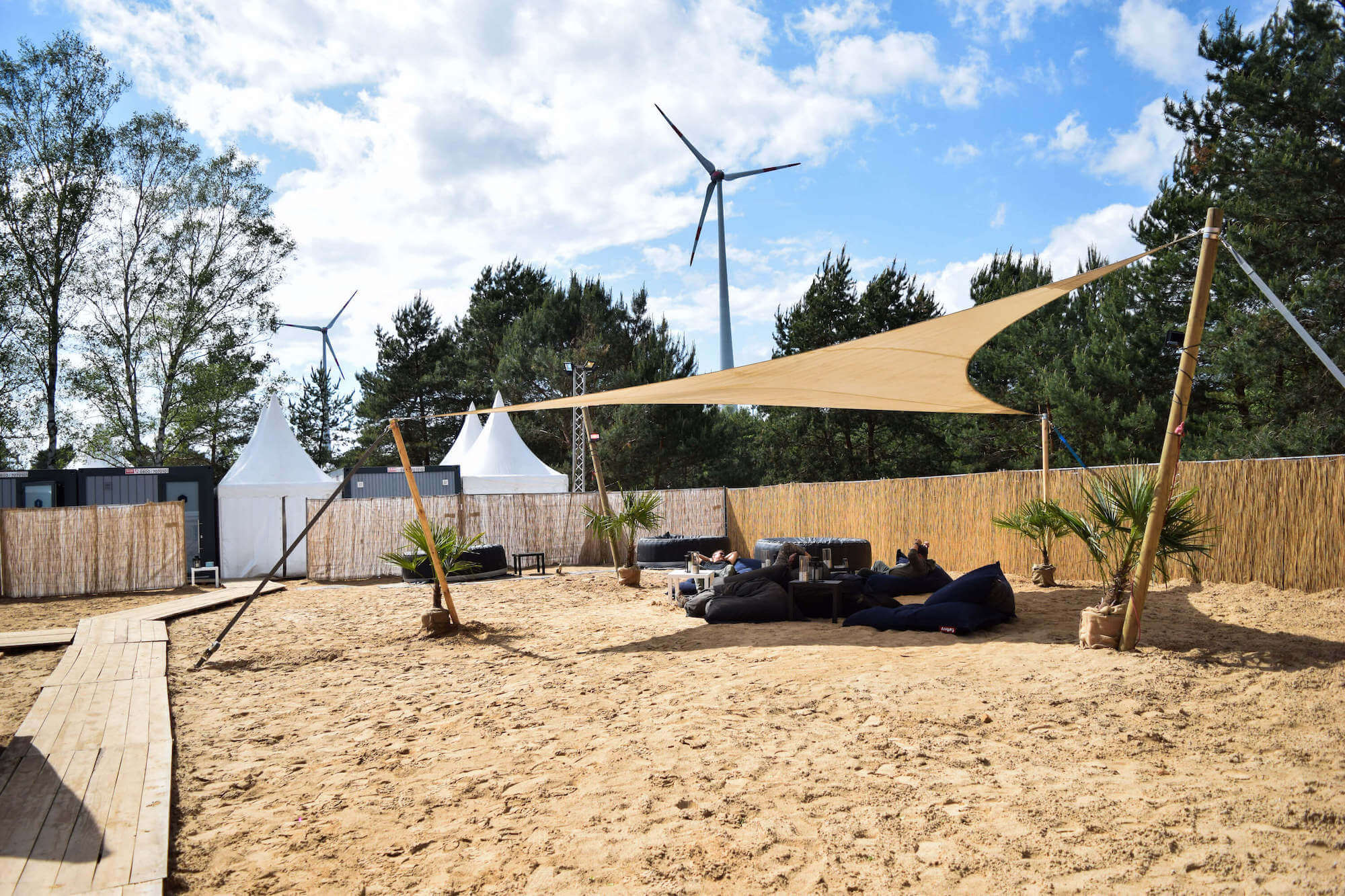 Sandige Outdoor-Lounge mit Sonnensegel, Sitzsäcken, Palmen und Zelten; Windrad im Hintergrund.