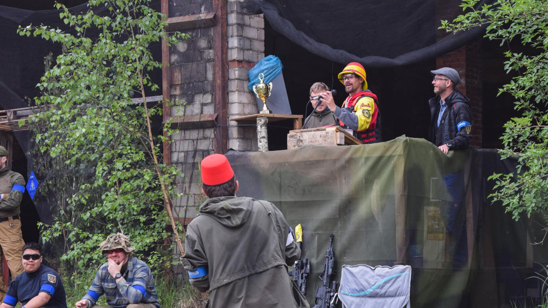 Airsoft-Veranstaltung mit Trophäe, Redner am Podium, Teilnehmer mit blauen Bändern, grüne Tarnung.