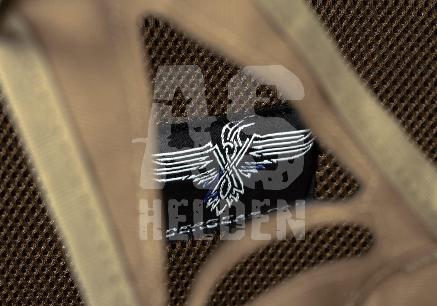 Schwarzes Patch mit Flügel-Emblem auf braunem, atmungsaktivem Stoff.