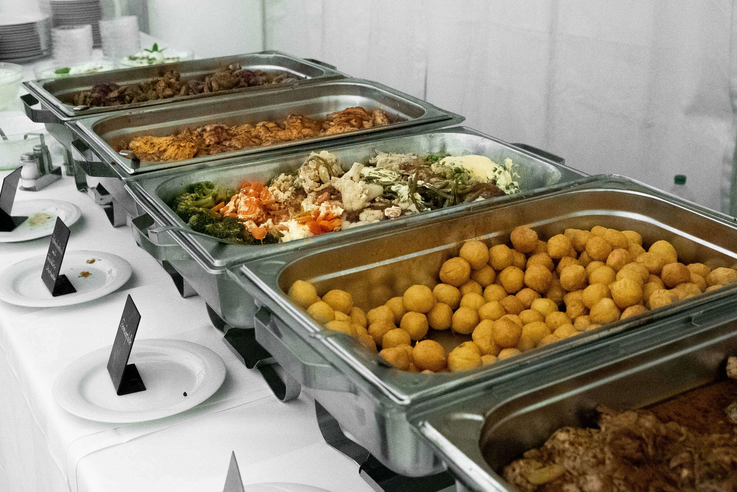 Buffet mit mehreren Edelstahlbehältern, gefüllt mit verschiedenen Speisen, auf einem weißen Tisch.