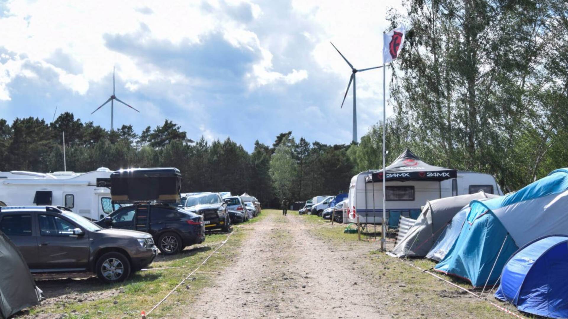 Campingplatz mit Zelten und Wohnmobilen entlang eines Wegs, im Hintergrund zwei Windräder.