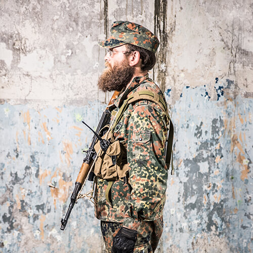 Person in Flecktarn-Uniform mit Gewehr im Profil vor einer rustikalen Wand.