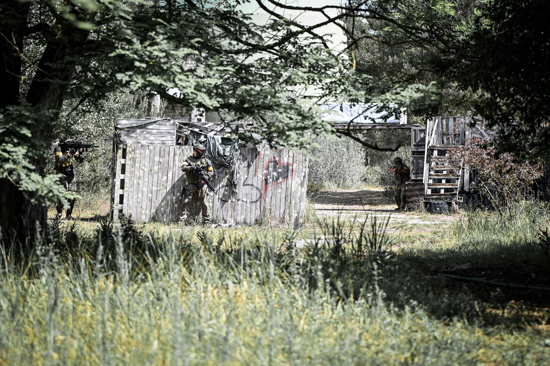 Airsoft-Spieler in Tarnkleidung manövrieren durch bewaldetes Gelände mit Holzhütte.