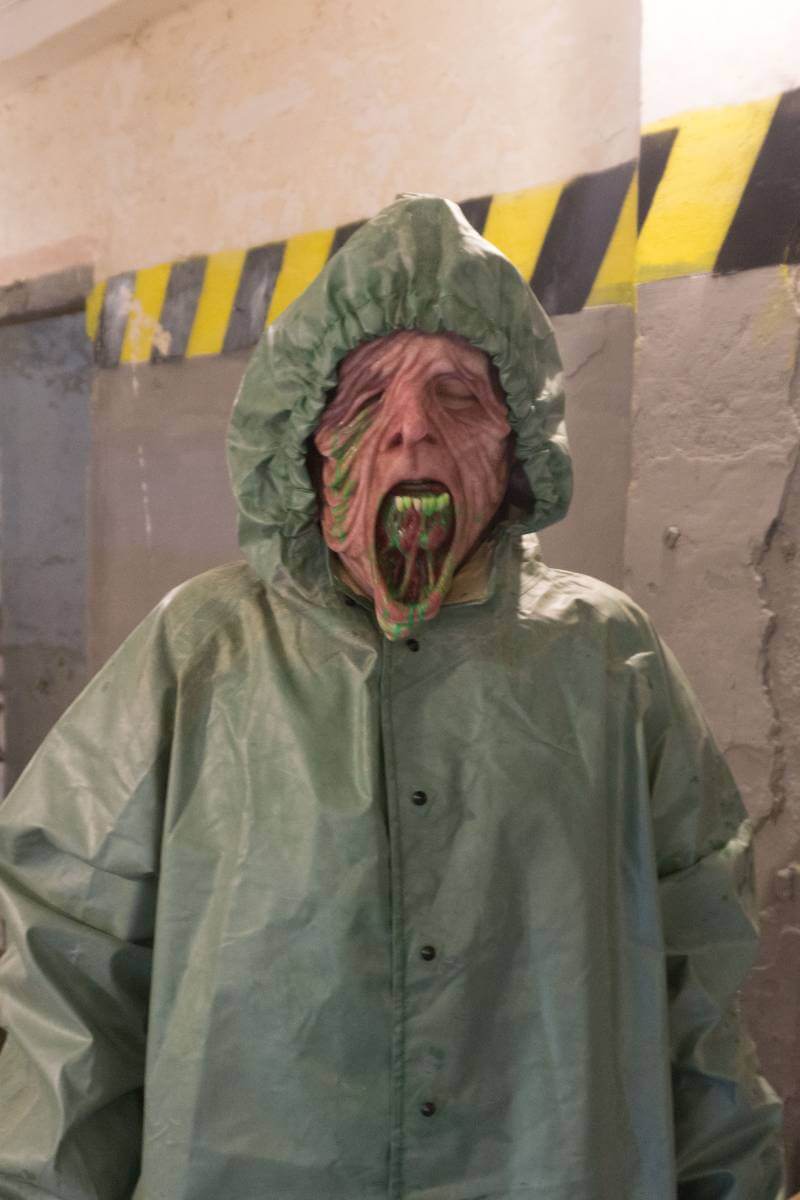 Person in grünem Schutzanzug mit grotesker Maske und weit geöffnetem Mund; Hintergrund mit gelb-schwarzen Markierungen.