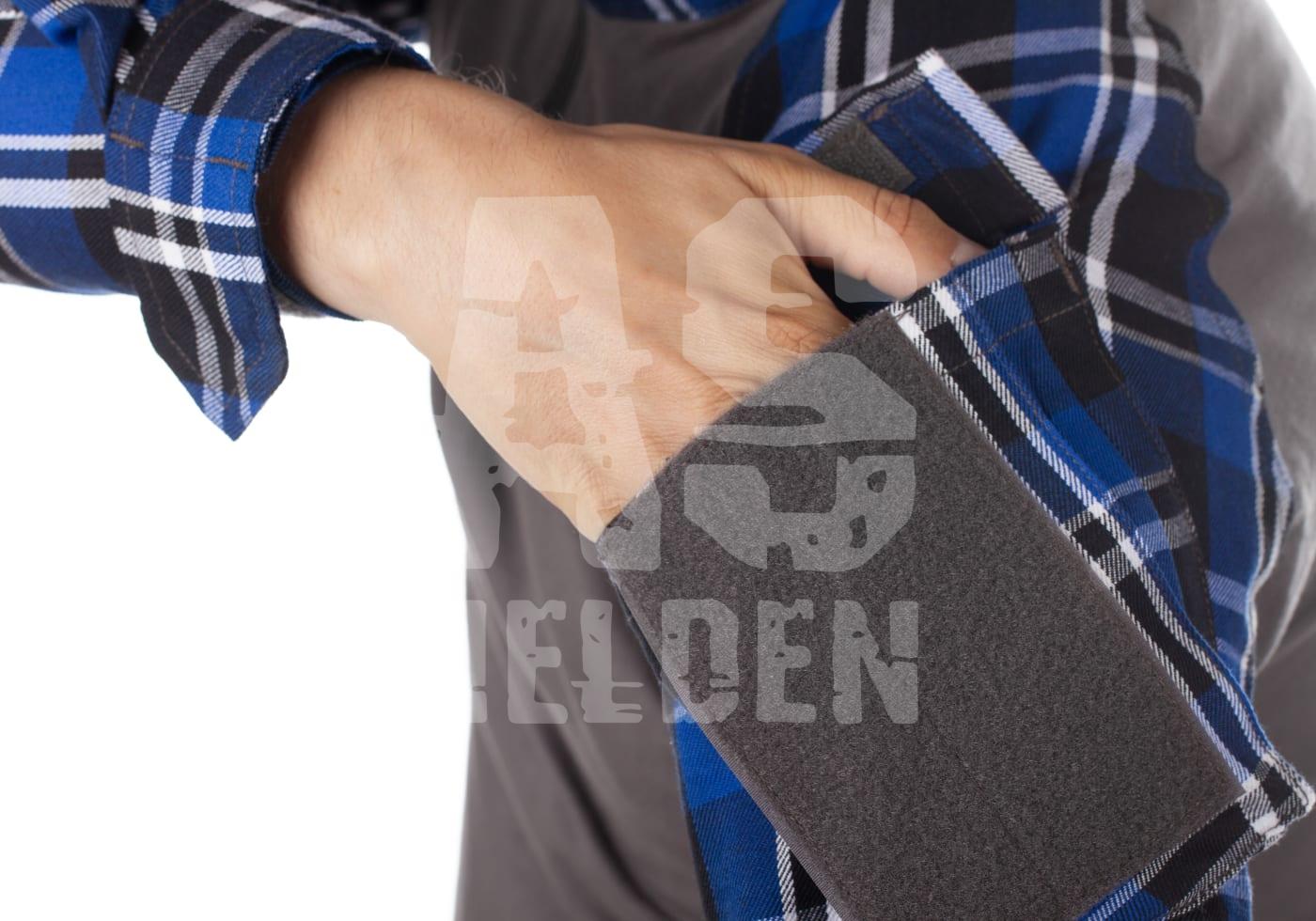 Flannel Combat Shirt Person steckt Hand in die abnehmbare Tasche eines blau karierten Hemds.