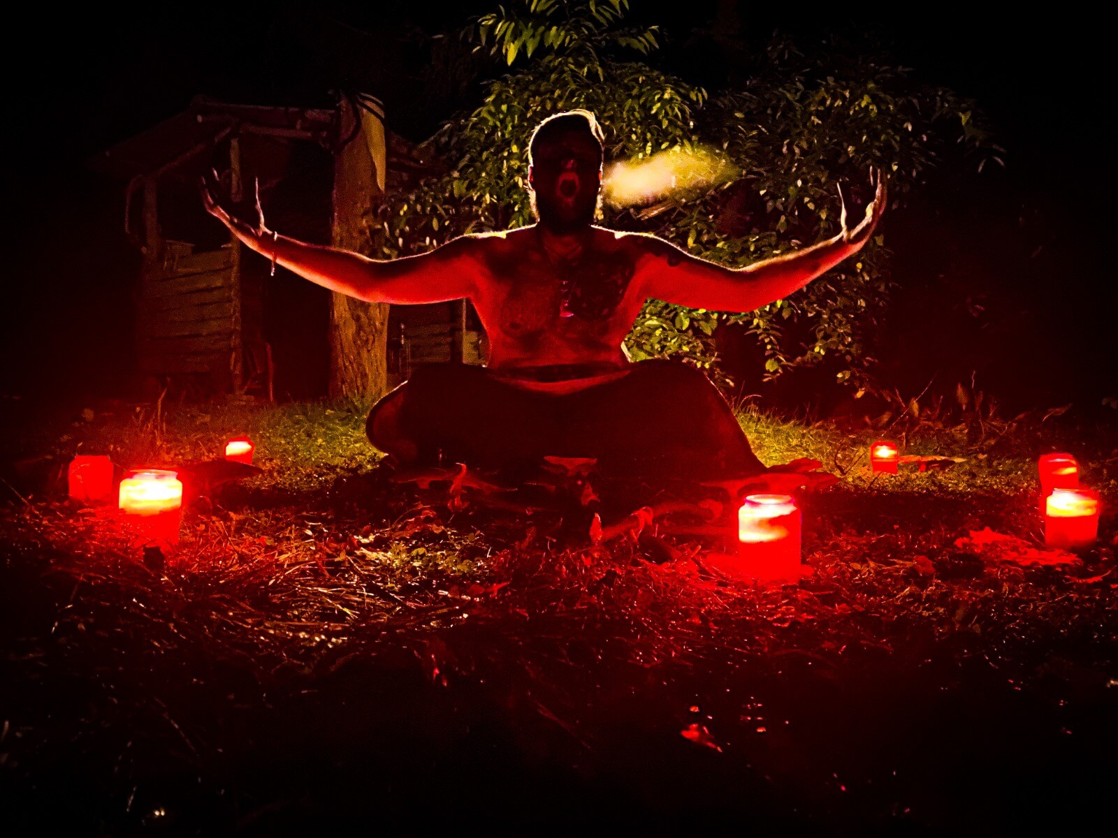 Person sitzt im Dunkeln auf Boden, umgeben von roten Kerzen, mit ausgestreckten Armen, wirkt meditativ.