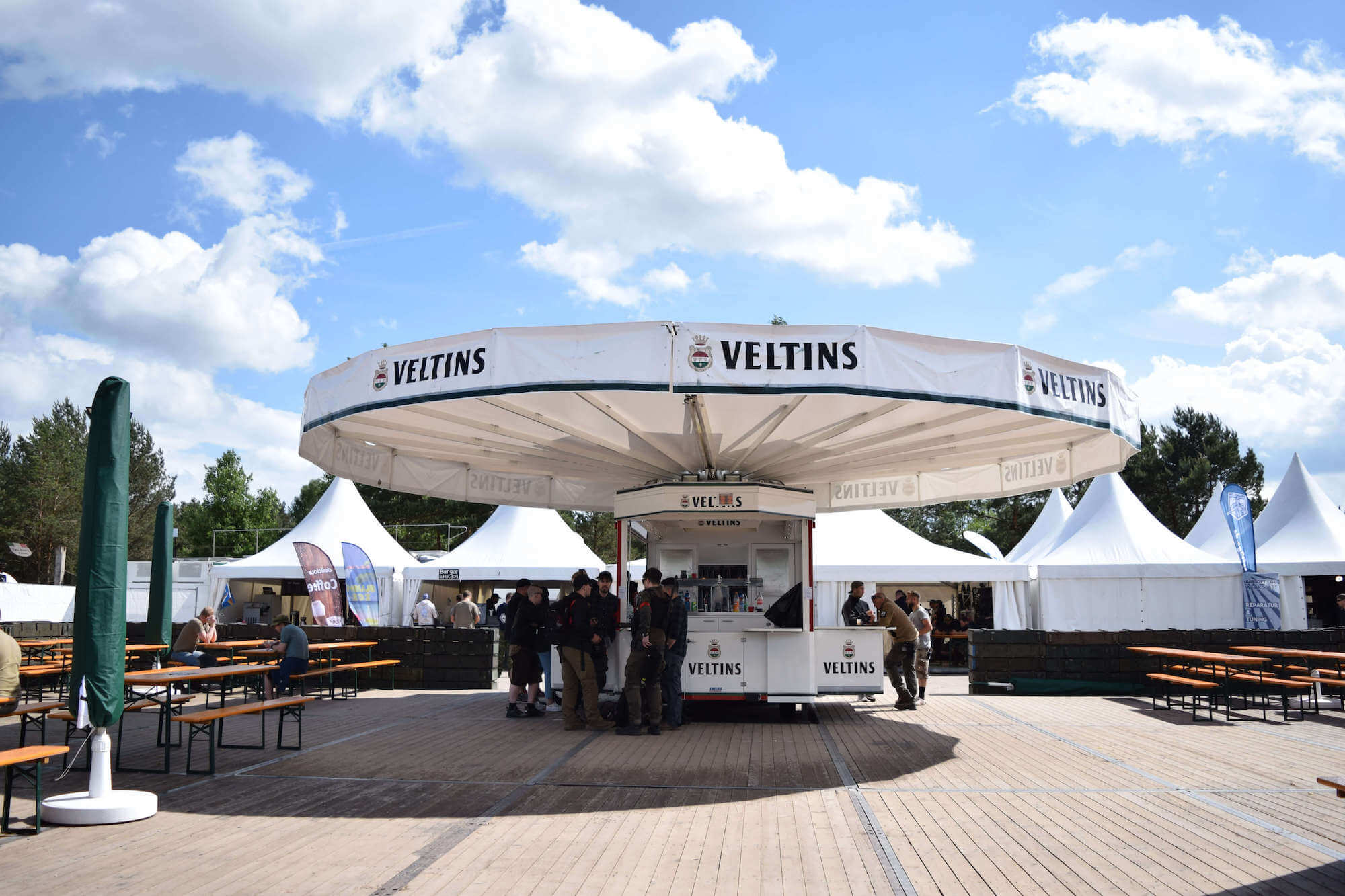 Großer Veltins-Stand auf einem Festivalgelände mit Biertischen, Zelten und Besuchern unter blauem Himmel.