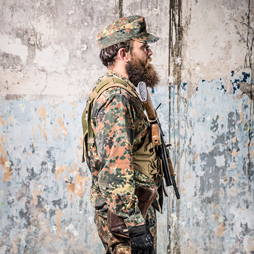 Mann in Tarnkleidung mit Airsoft-Gewehr und Mütze vor urbaner Wand.