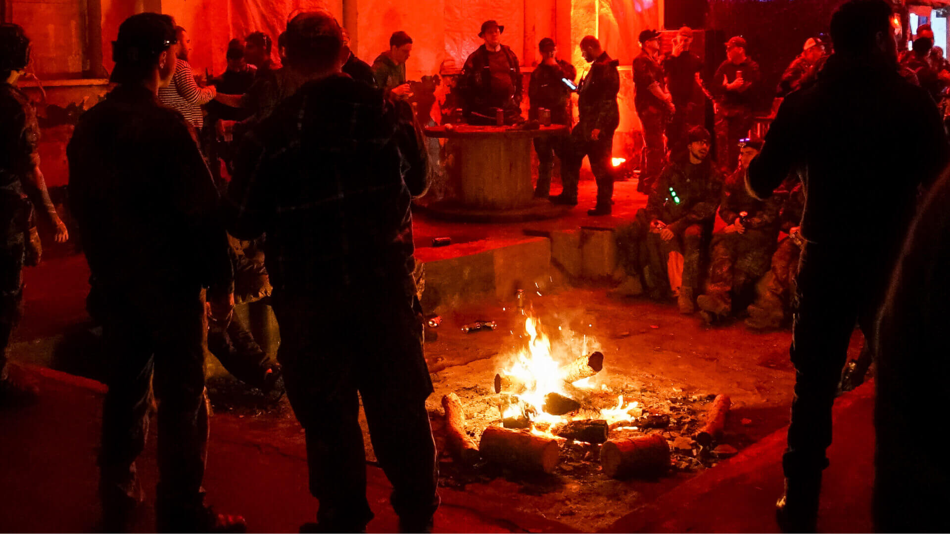 Airsoft-Spieler versammeln sich um ein Lagerfeuer bei rotem Licht in einer entspannenden Atmosphäre.