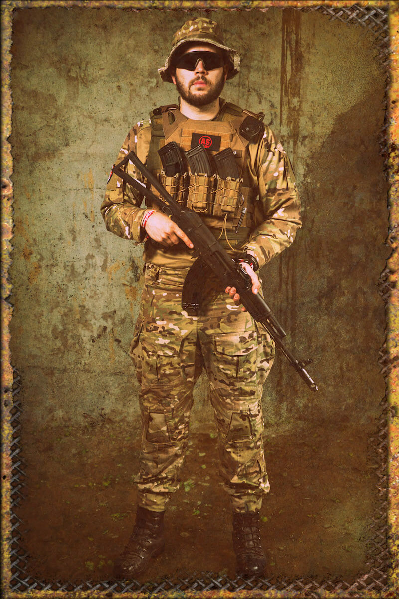 Airsoft-Spieler in Tarnkleidung hält Gewehr, trägt Schutzbrille und Hut, vor rustikalem Hintergrund.