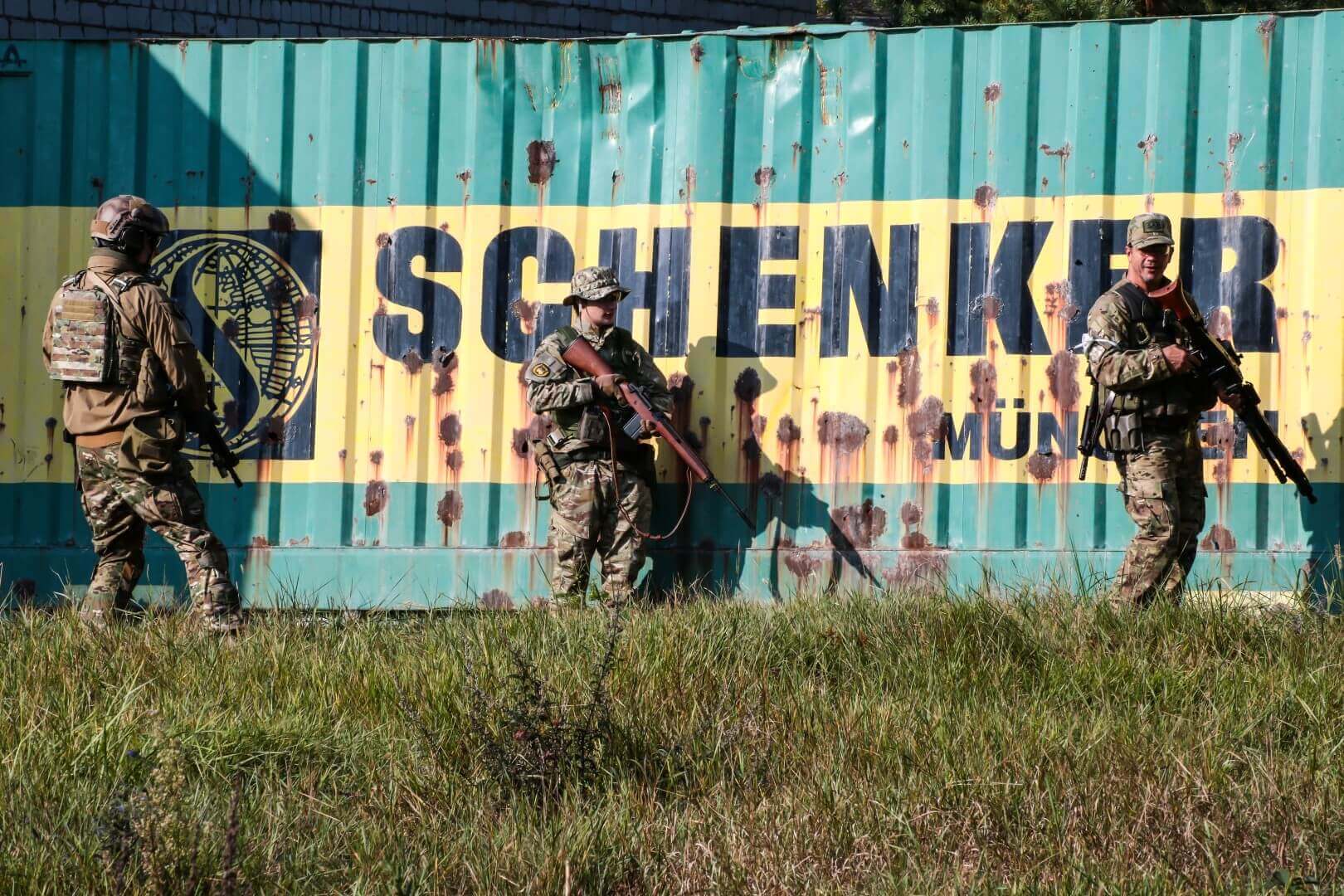 Drei Airsoft-Spieler in Tarnkleidung vor einem Container mit "Schenker"-Logo im Hintergrund.