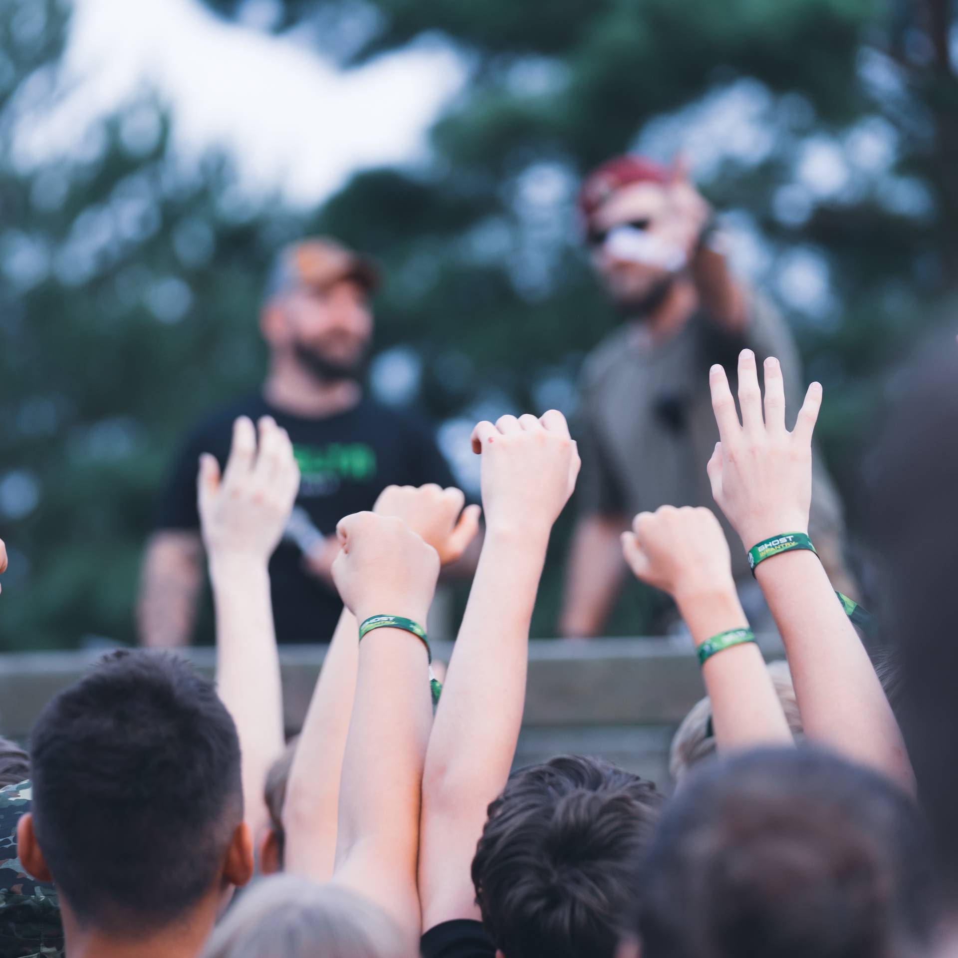 Airsoft-Treffen, Teilnehmer heben Arme mit grünen Armbändern, unscharfer Hintergrund mit zwei Personen.