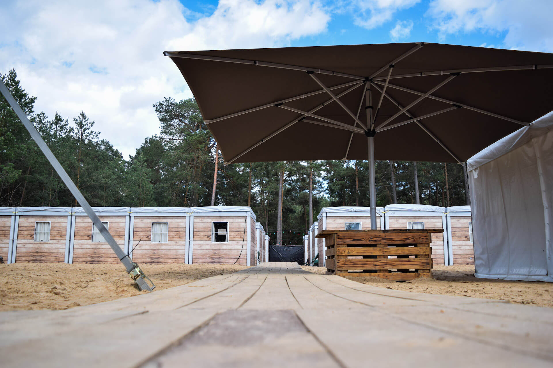 Sonnenschirm über Holztischen vor Containergebäuden im Wald, Airsoft-Basislager-Setup.
