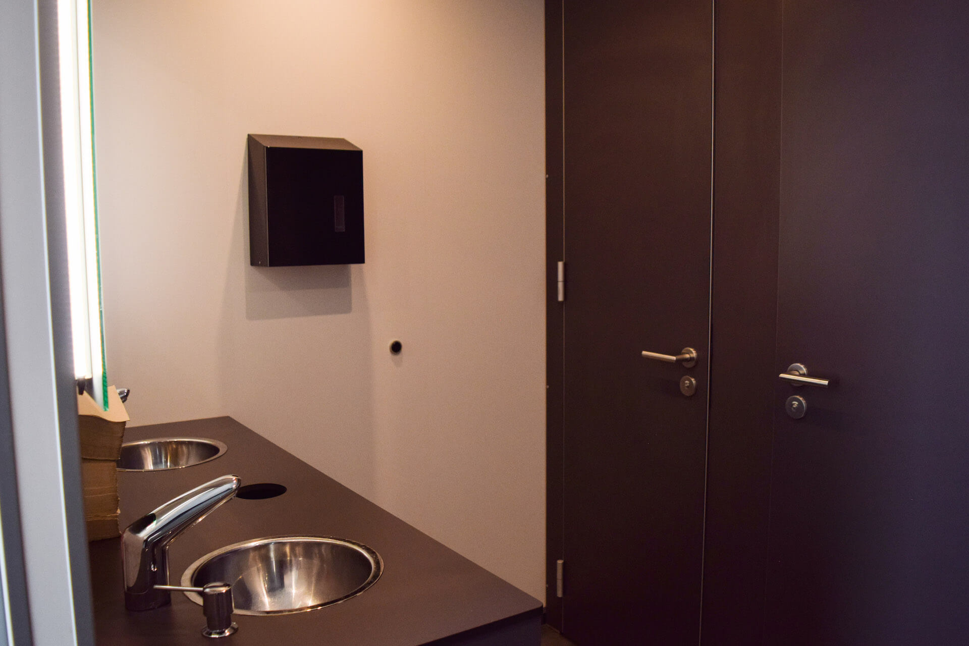 Zwei Waschbecken mit Wasserhähnen in einem modernen Badezimmer, links ein Papierhandtuchspender, rechts Türen.