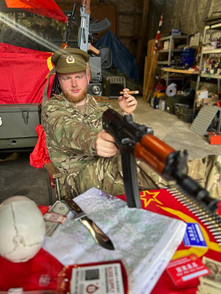 Person in Tarnkleidung sitzt entspannt mit Airsoft-Waffe, Tabak und Landkarte, umgeben von roten Flaggen und Zubehör.