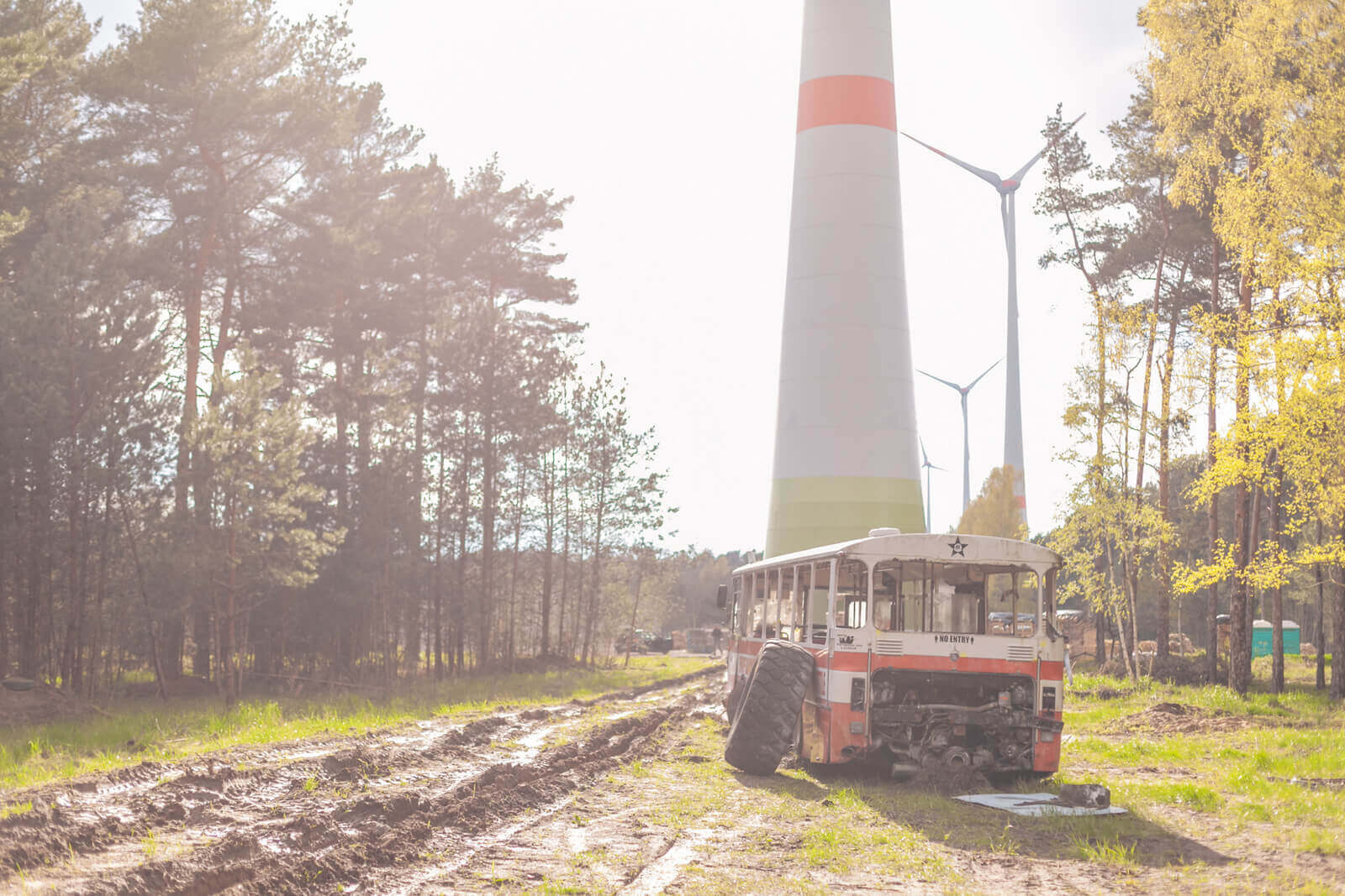 Verlassener Bus vor Windkraftanlagen und Wald auf schlammigem Weg.