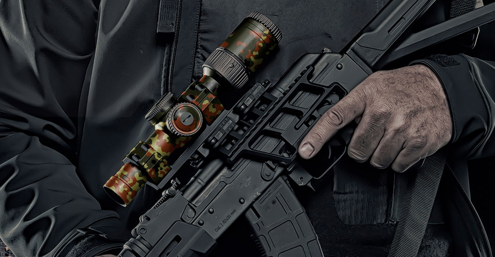 Person hält Airsoft-Gewehr mit Camouflage-Zielfernrohr, trägt schwarze Jacke.