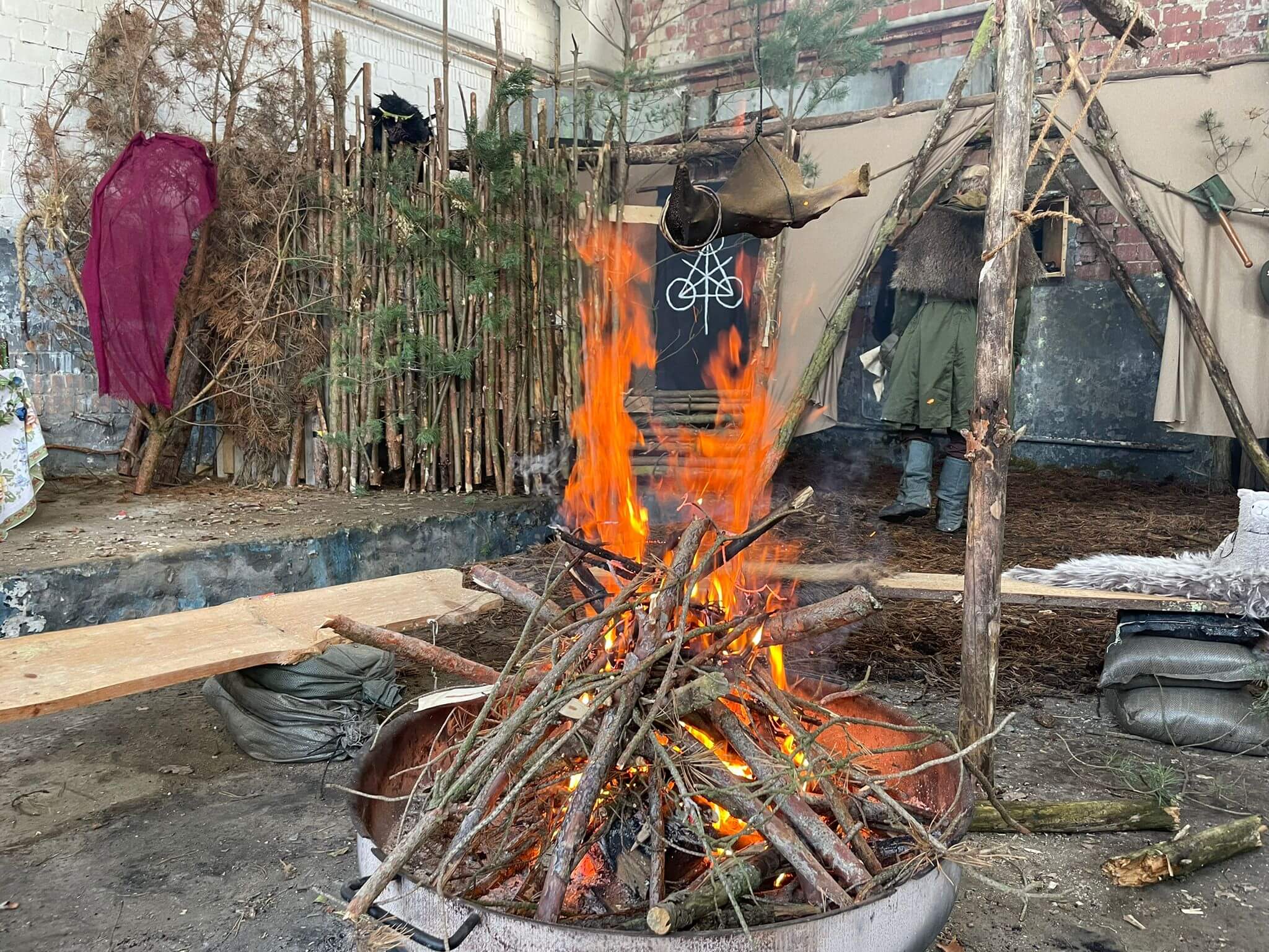 Lagerfeuer in rustikaler Indoor-Szene mit Holzhütte und Tarnmaterialien.