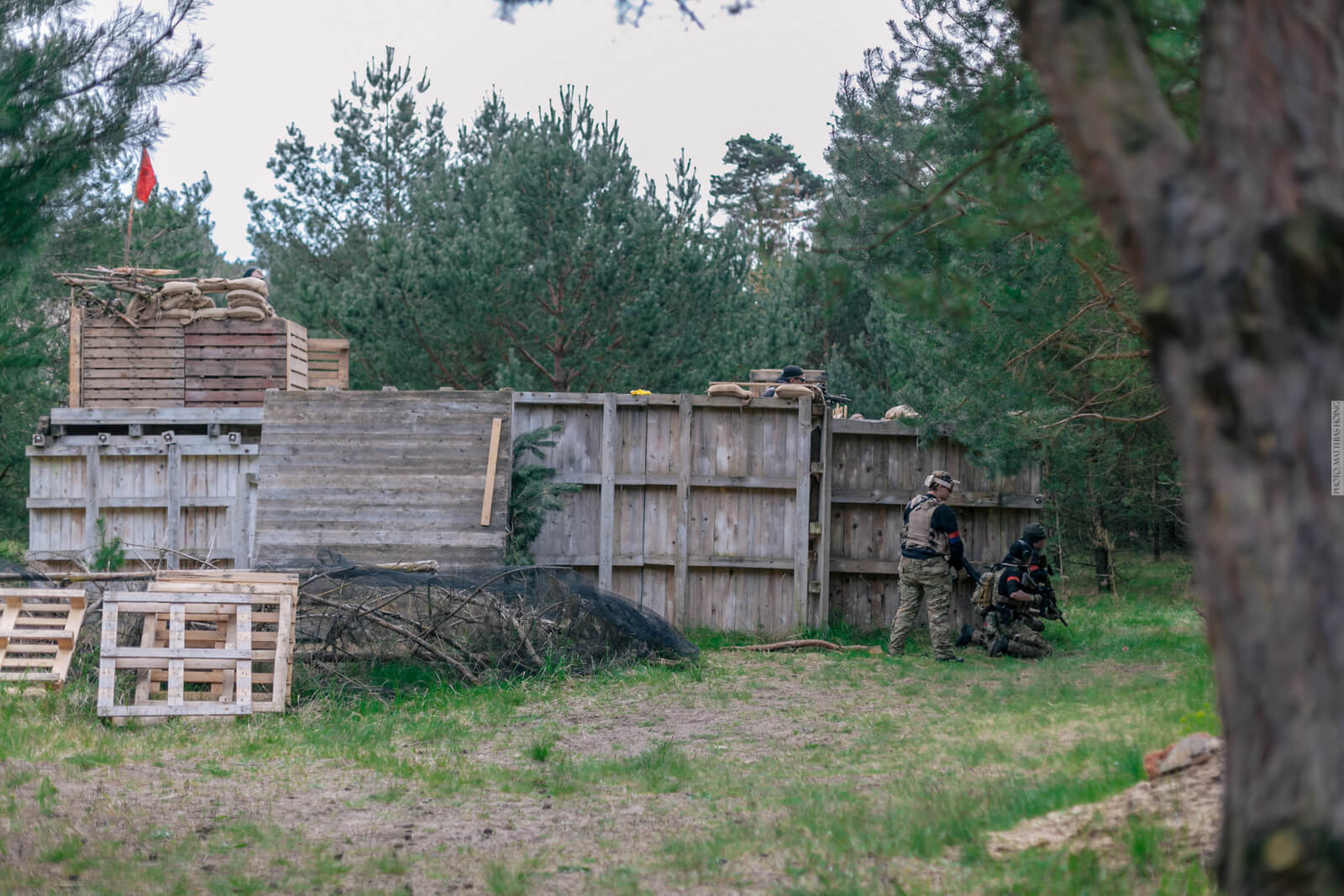 Airsoft-Spieler in Tarnkleidung an hölzerner Festung im Wald, rote Flagge auf Wachturm.