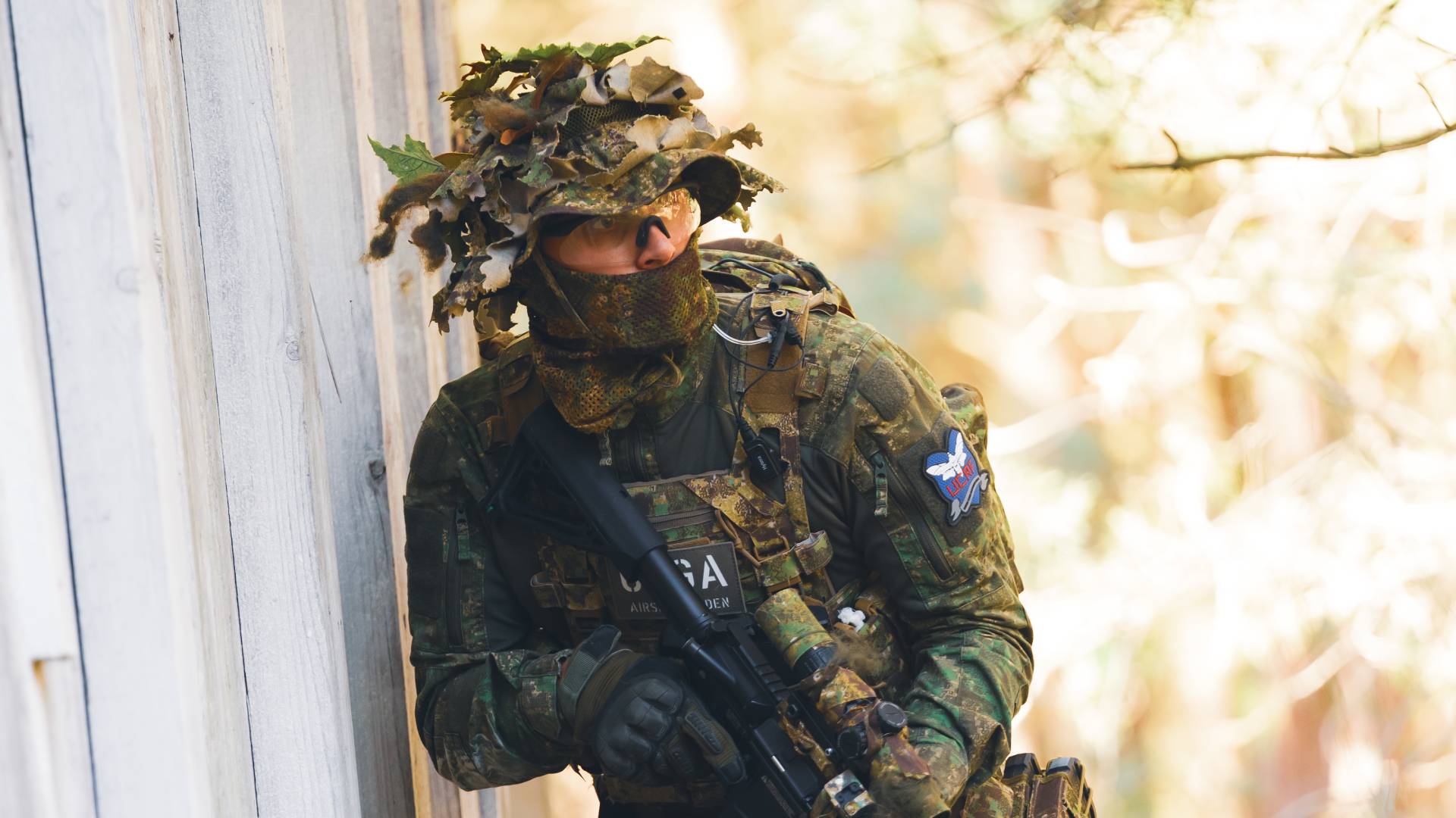 Airsoft-Spieler in Tarnkleidung lehnt an Wand, hält ein Gewehr, bereit für den Einsatz im Gelände.