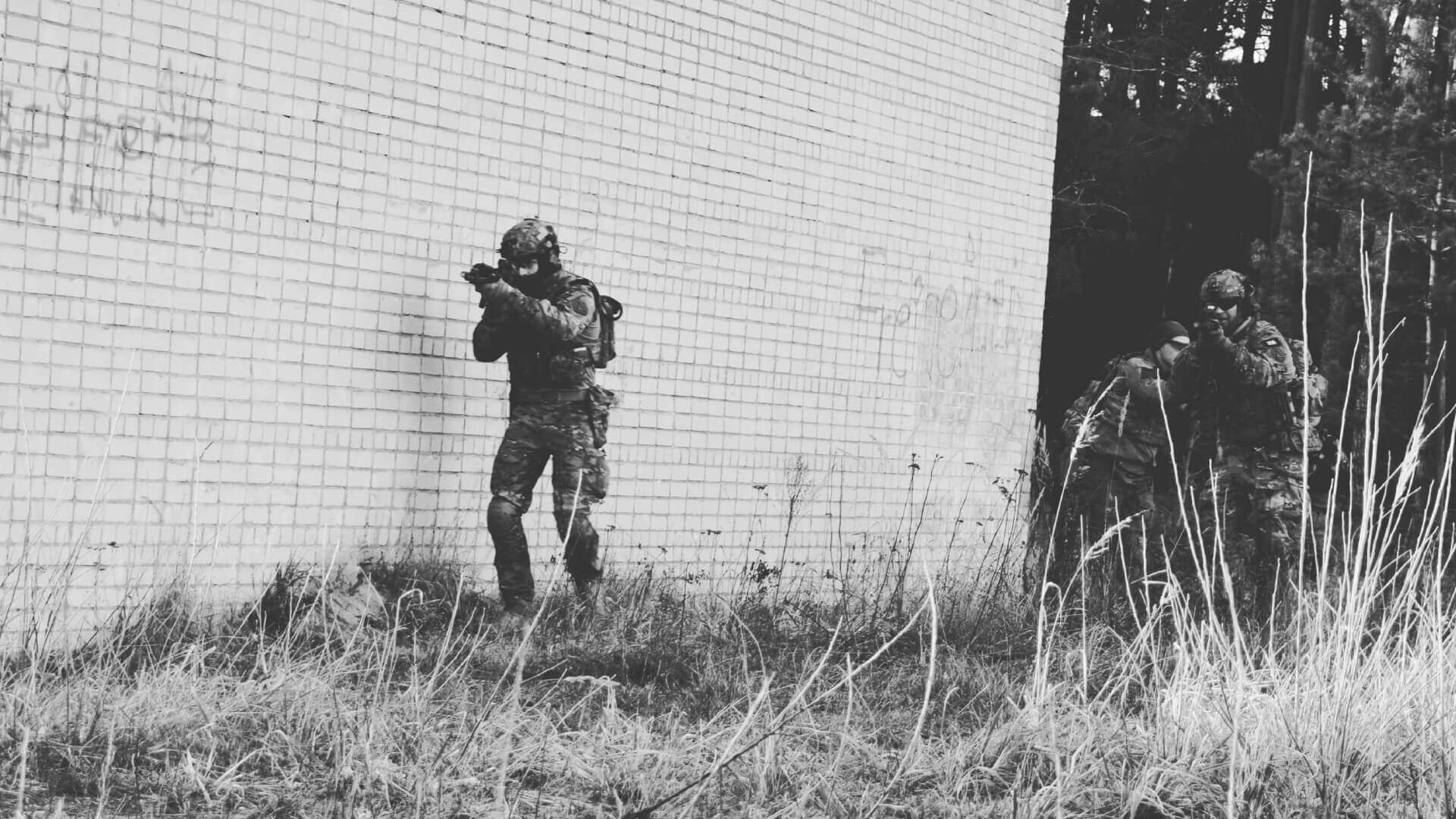 Airsoft-Spieler in Tarnkleidung, sichern ein Gebäude mit Schusswaffen. Draußen, neben einer Mauer.