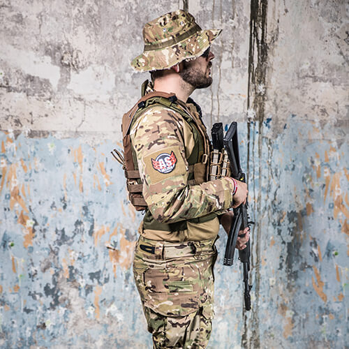 Person in Camouflage mit Hut, Weste und Airsoft-Gewehr vor verwitterter Wand.