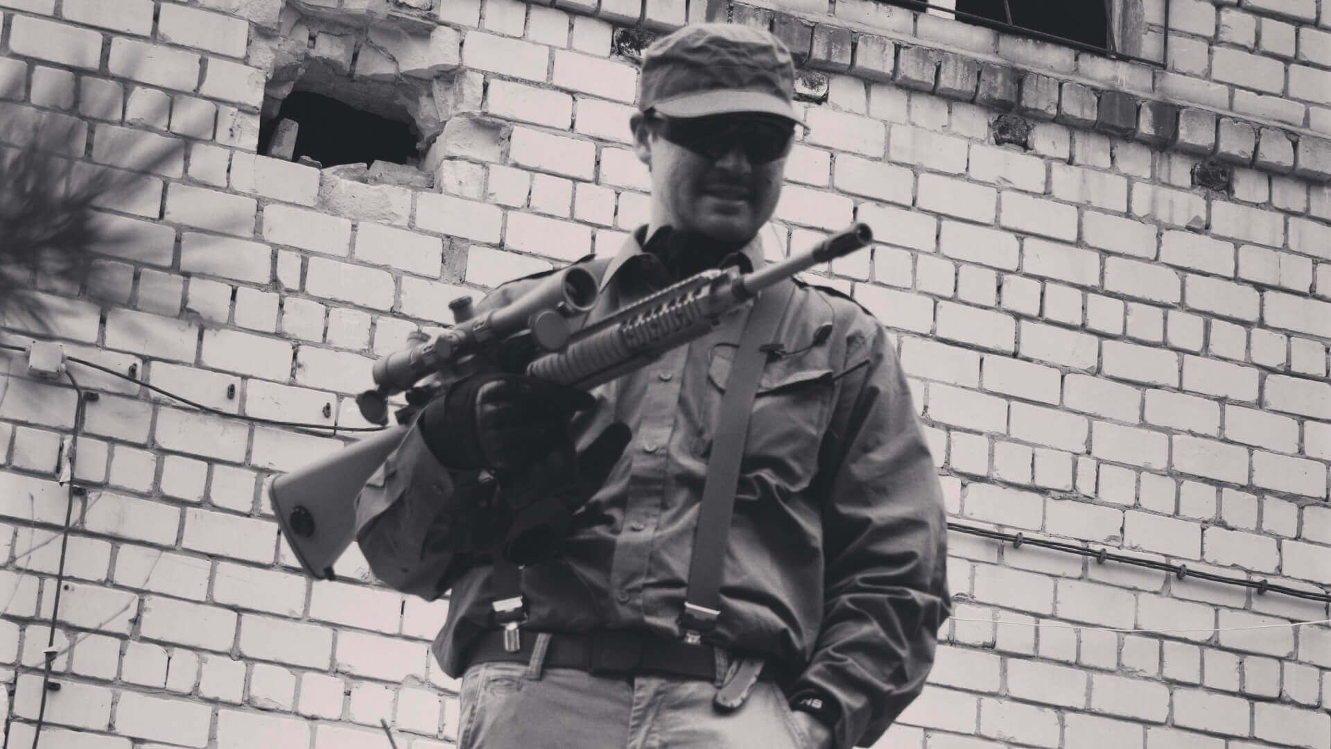 Person hält Gewehr vor beschädigter Ziegelwand, trägt Militärkleidung und Mütze. Schwarz-Weiß-Foto.