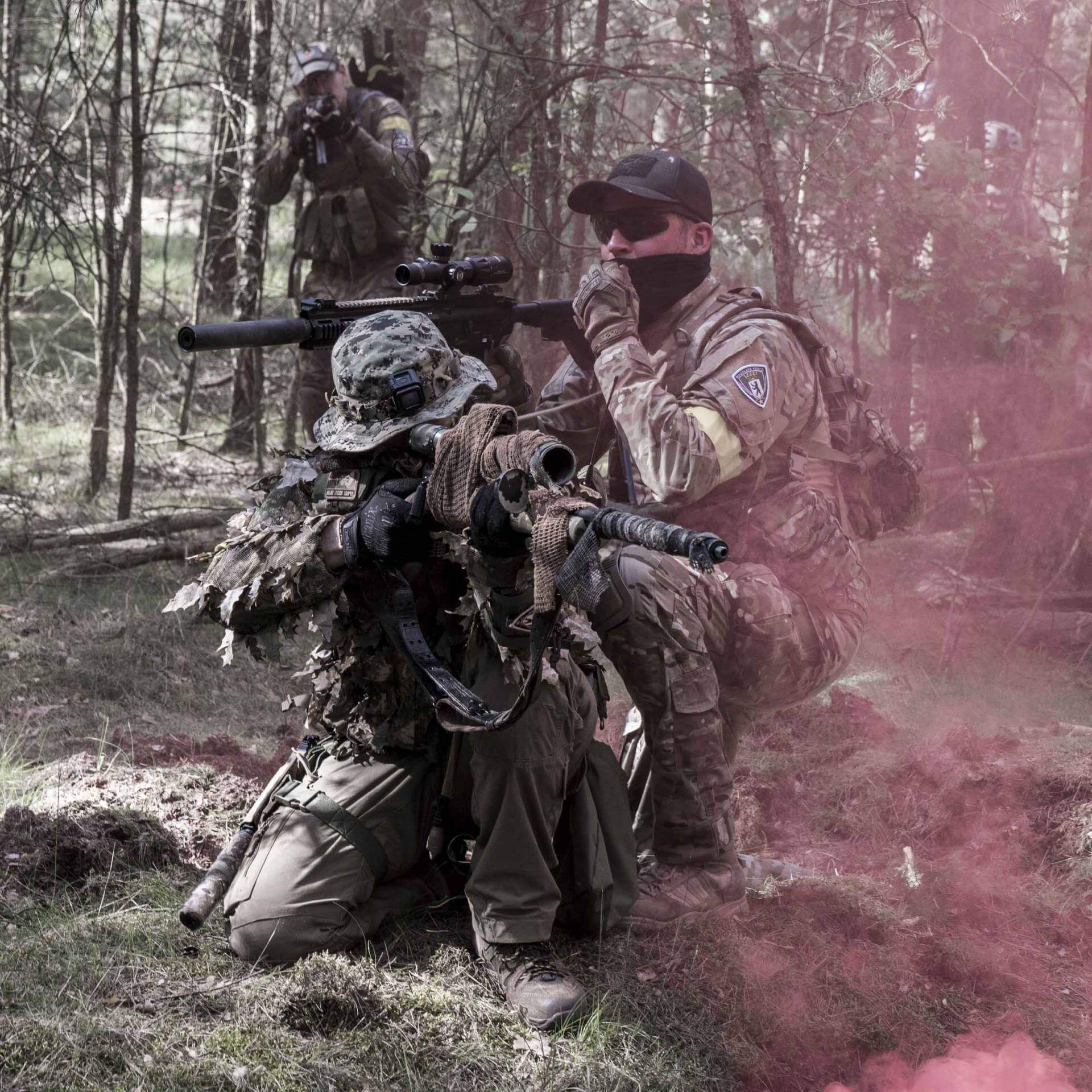 Airsoft-Spieler im Wald mit Scrimghillie, Gewehr und rotem Rauch. Taktische Ausrüstung im Einsatz.