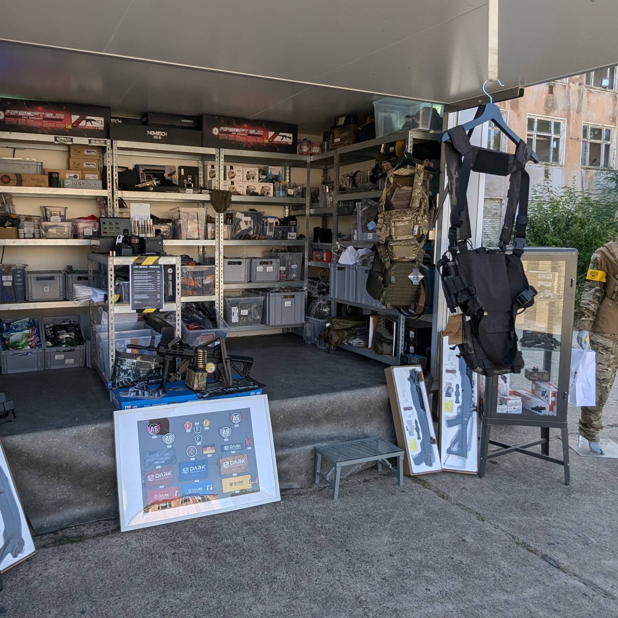 Airsoft-Stand mit Zubehör, taktischen Westen und Ausrüstung, auf einem Tisch und Regalen ausgestellt.