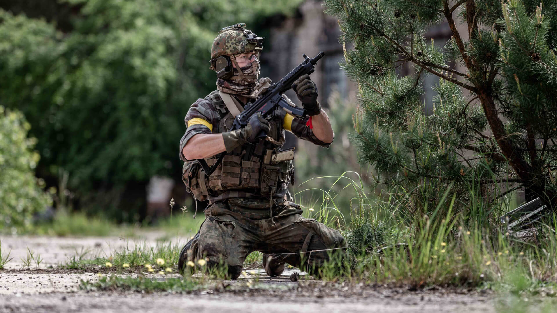 Airsoft-Spieler in Tarnkleidung, kniend mit Gewehr im Anschlag, in grüner Waldumgebung.
