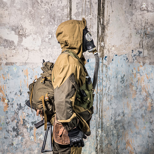 Person in taktischer Ausrüstung mit Gasmaske und Rucksack vor verwitterter Wand.