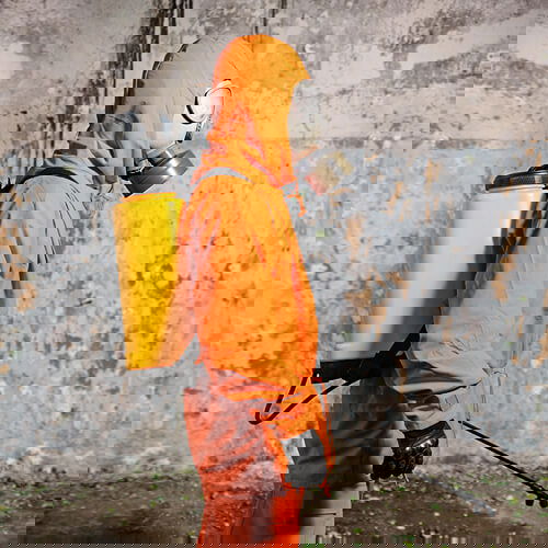 Person in orange Schutzanzug mit Gasmaske und Sprühtank, vor betonierter Wand.