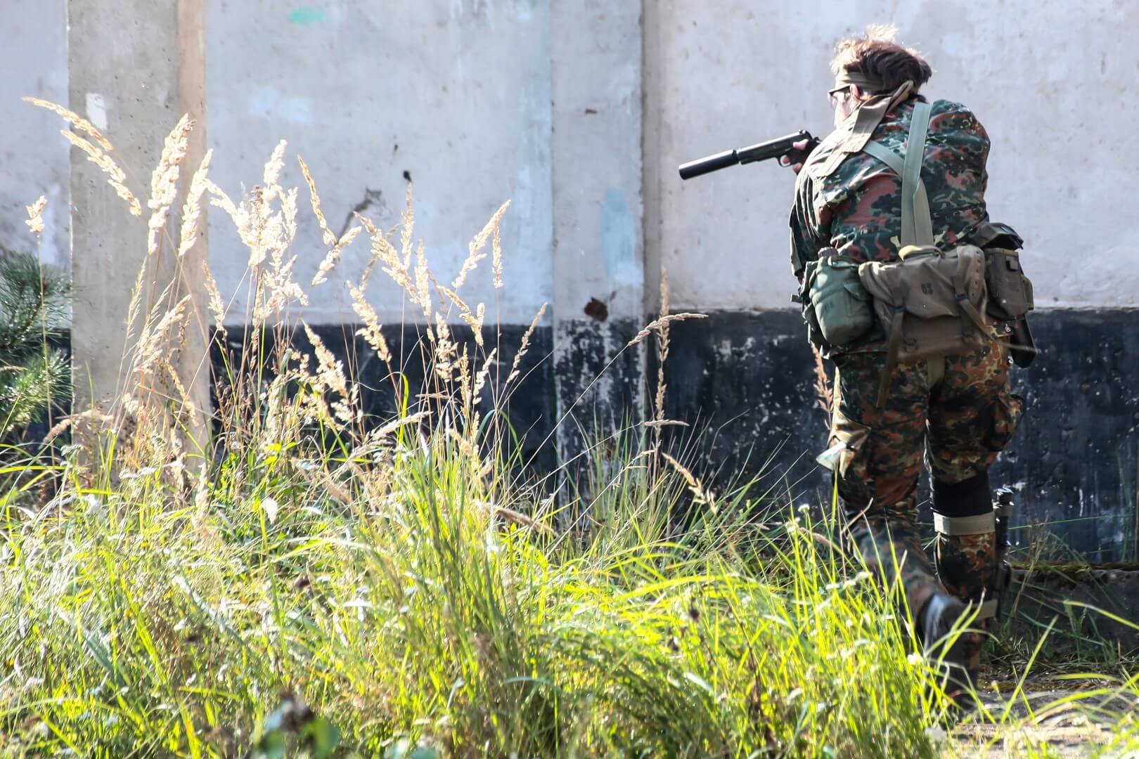 Person in Tarnkleidung mit Airsoft-Gewehr hinter Gras in urbaner Umgebung.