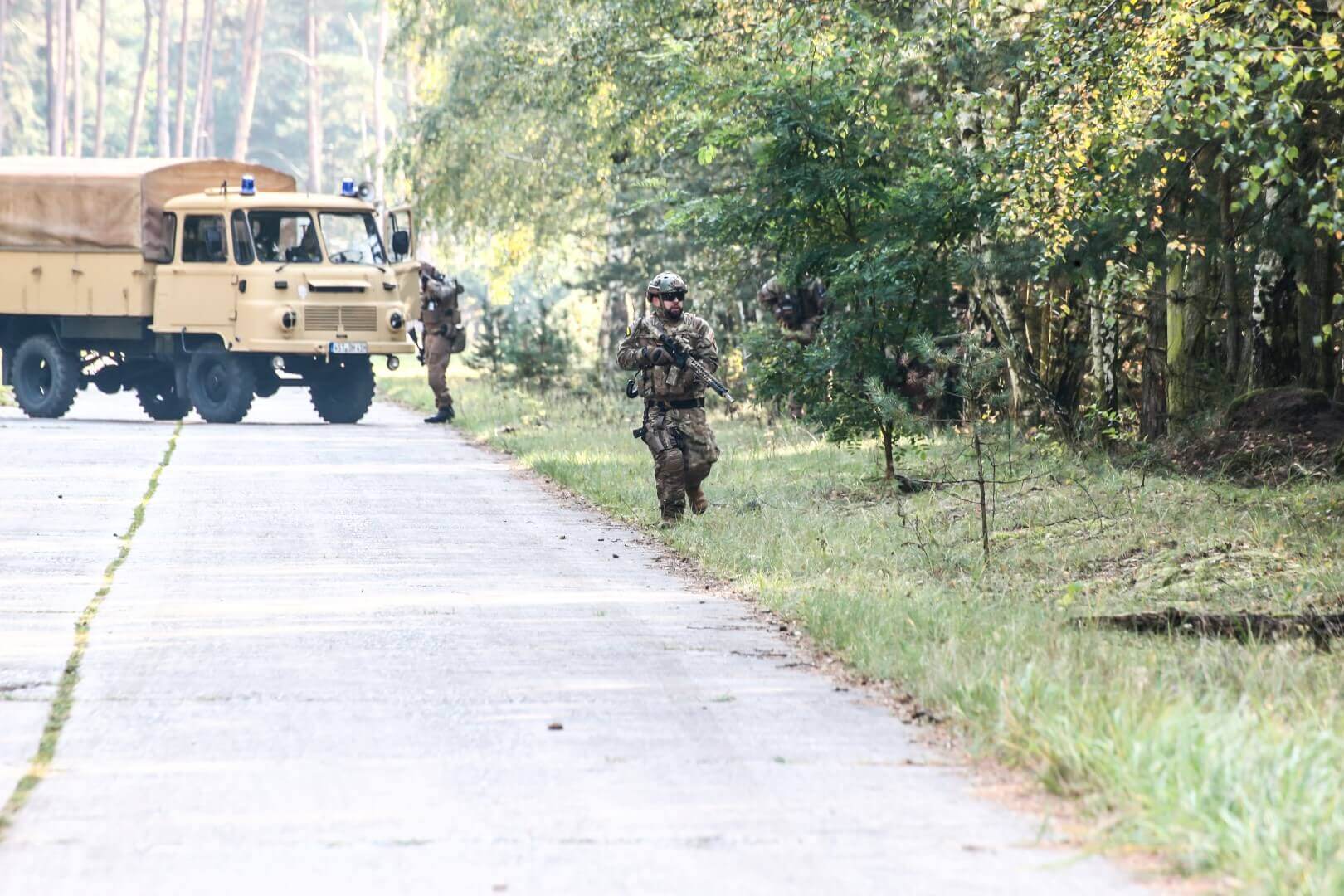 Airsoft-Spieler mit Ausrüstung neben Waldweg, Militärfahrzeug im Hintergrund.