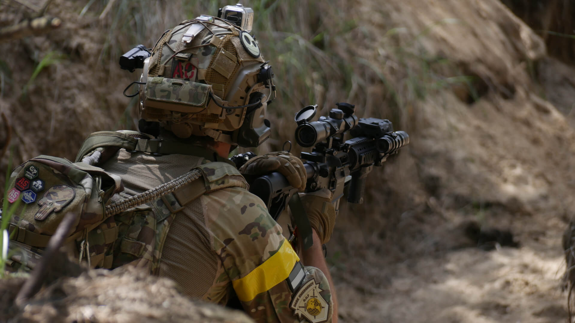 Airsoft-Spieler in Tarnkleidung, Helmkamera, gelbem Armband, mit Gewehr in Deckung im Gelände.