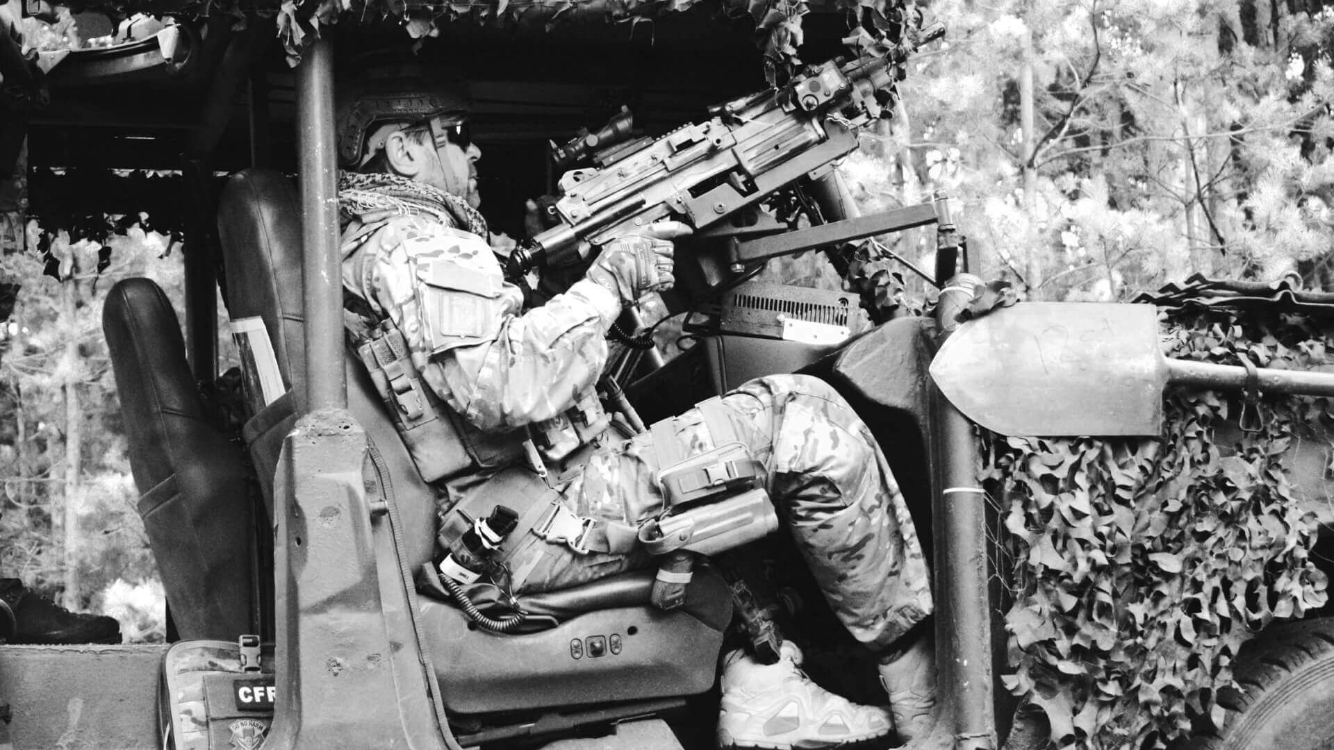 Airsoft-Spieler in voller Ausrüstung zielt aus einem getarnten Fahrzeug mit Gewehr.