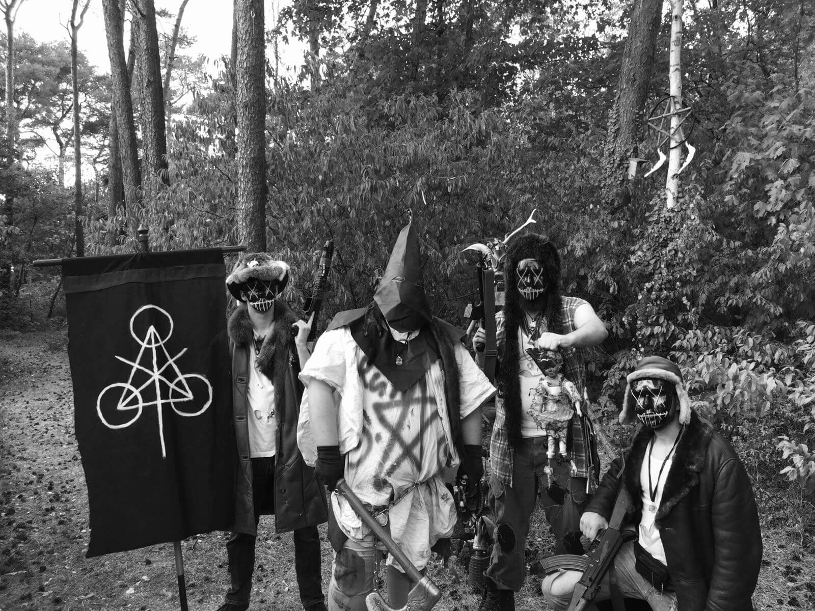 Vier maskierte Airsoft-Spieler im Wald mit Flagge und Ausrüstung, in post-apokalyptischer Kleidung.