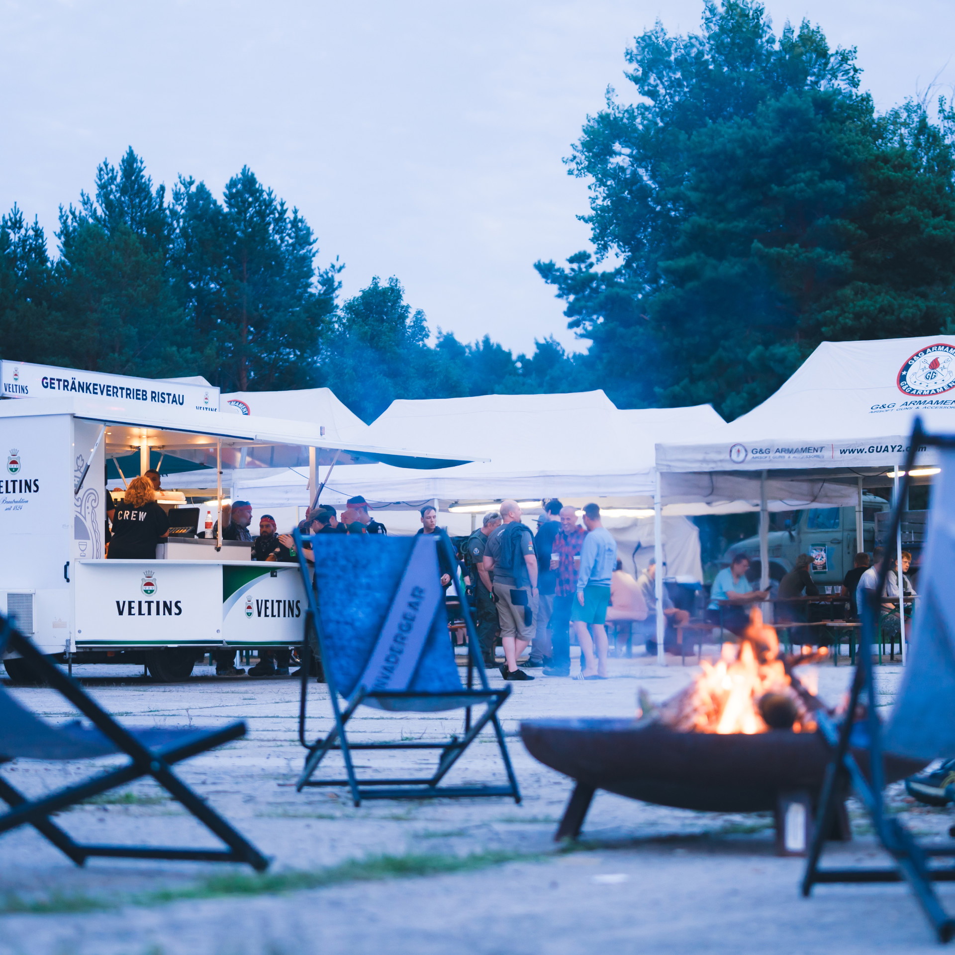 Grillfeuer und Getränkewagen auf Airsoft-Event, Teilnehmer entspannen in Camp-Stühlen.