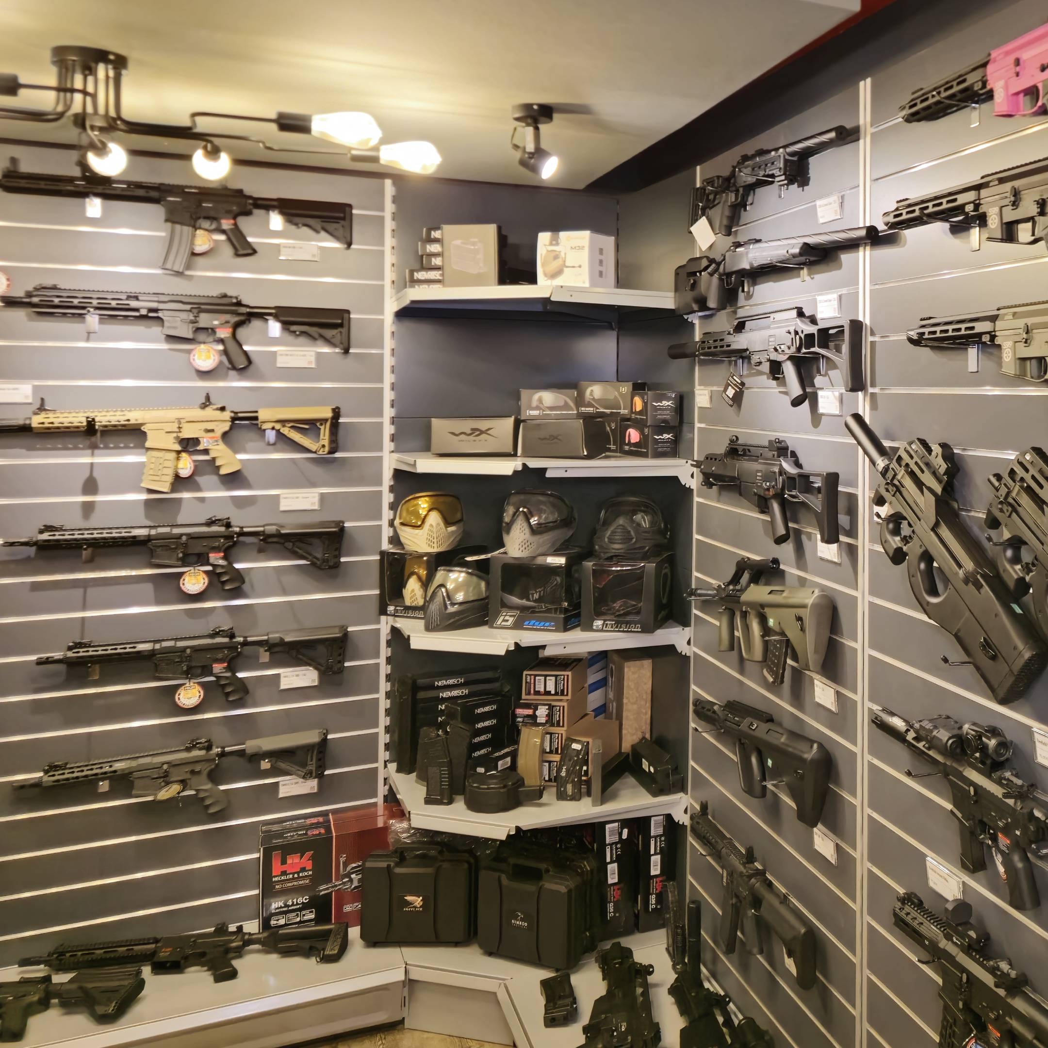 Airsoft-Schaufenster mit Gewehren, Pistolen, Masken und Zubehör an grauen Wänden ausgestellt.