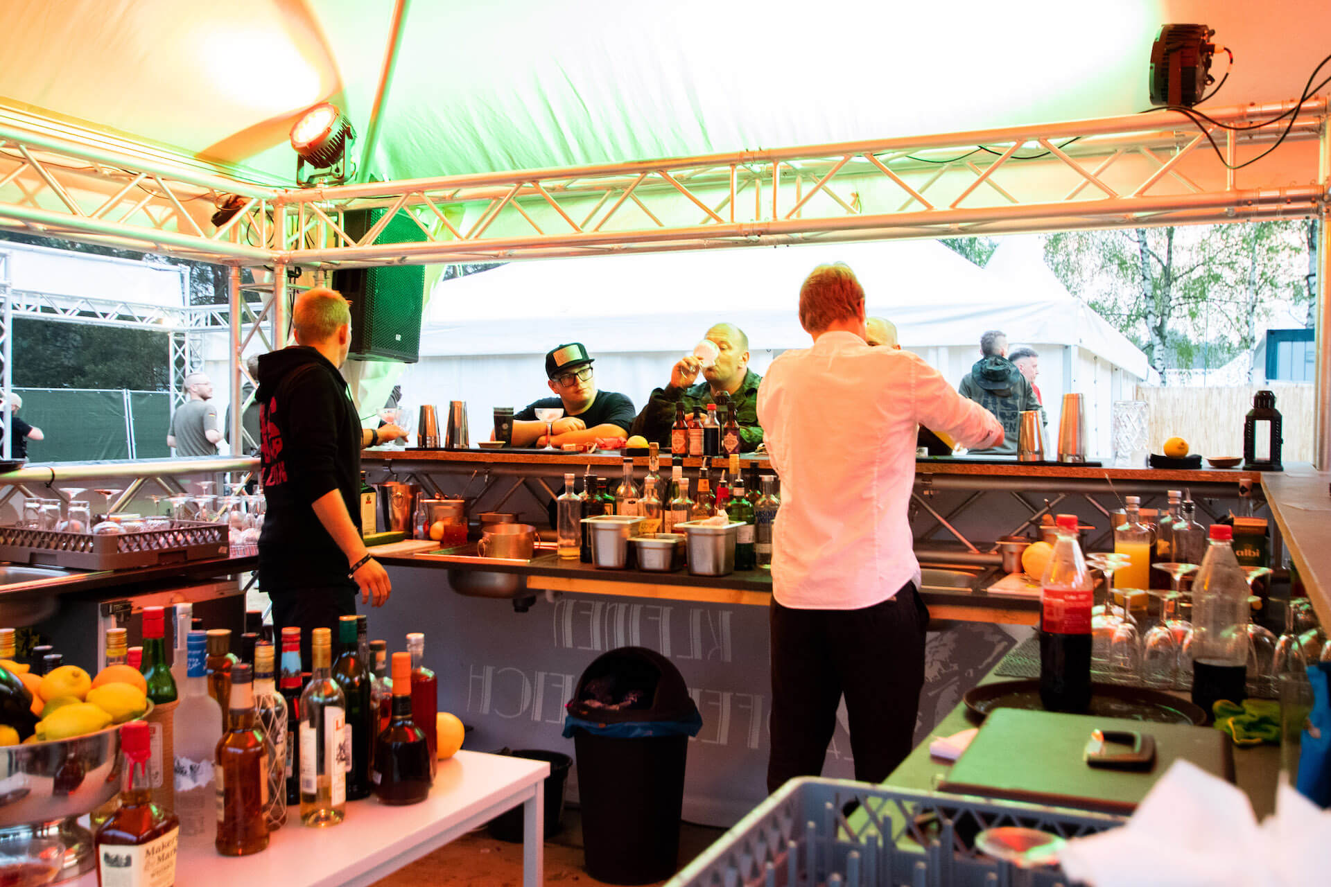 Barmänner mixen Getränke in einer Outdoor-Bar unter einem Zelt, während Gäste zuschauen und trinken.